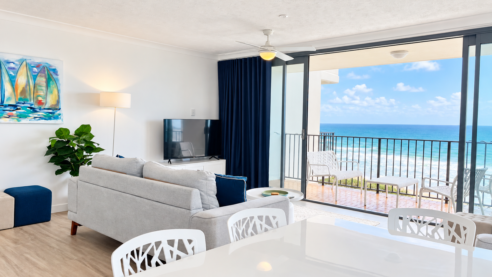 Modern living room with light gray sofa, blue pillows, a TV, indoor plant, and sailboat painting, opening to a balcony overlooking the ocean with white patio chairs and railing under a blue sky with clouds.