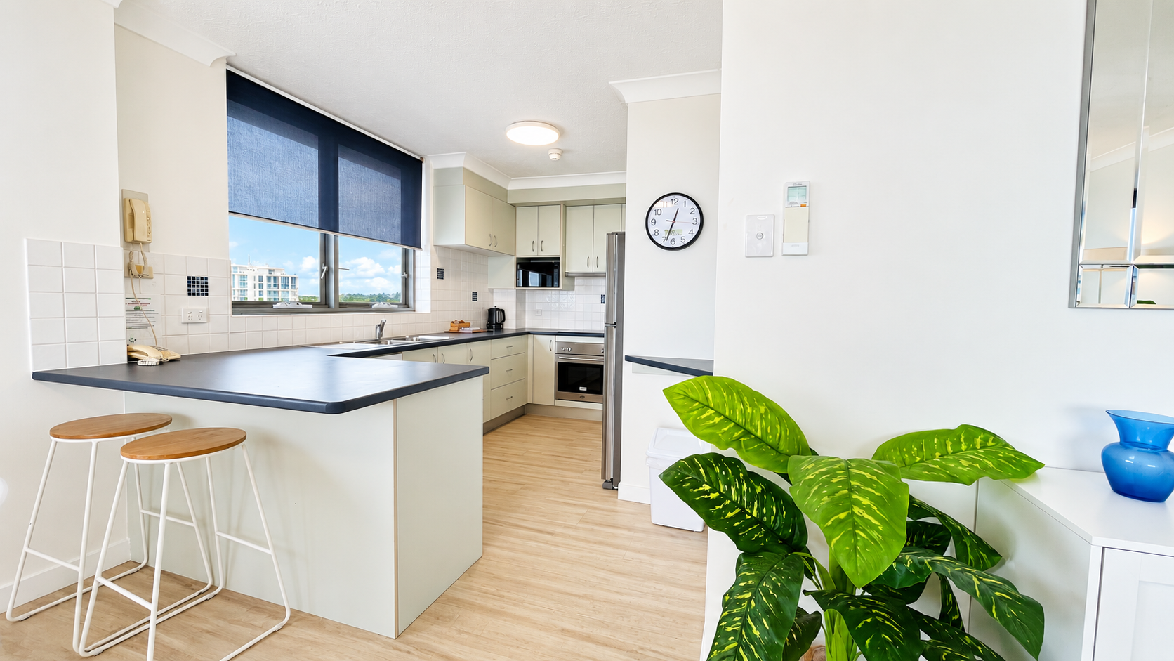 Bright modern kitchen with light wood flooring, white cabinets, black countertops, a window with a dark blue roller blind, and stainless steel appliances including a microwave and oven. There are two wooden stools with white metal legs at the counter, a wall clock showing 7:06, a green leafy plant near a white cabinet with a blue vase, and an intercom phone on the wall beside the window.