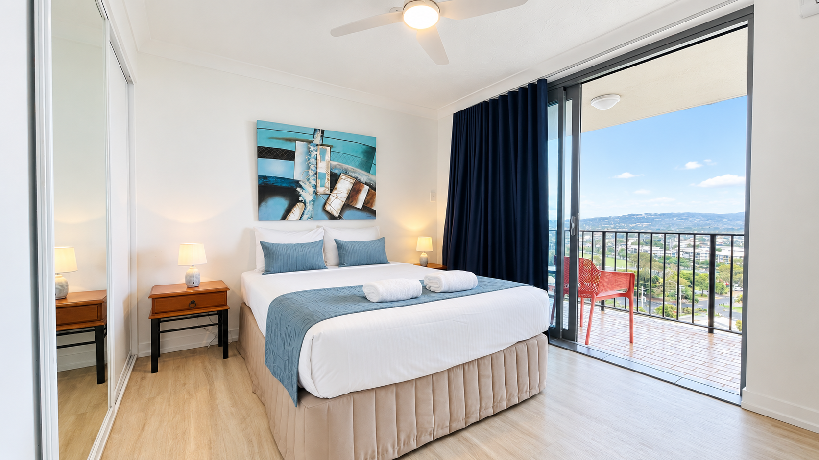 Modern bedroom with a double bed dressed in white linens and blue decorative pillows and runner, two rolled white towels placed on top, wooden nightstand with lit lamp, large sliding glass door leading to balcony with a red chair, and a scenic view of mountains and cityscape under a blue sky.