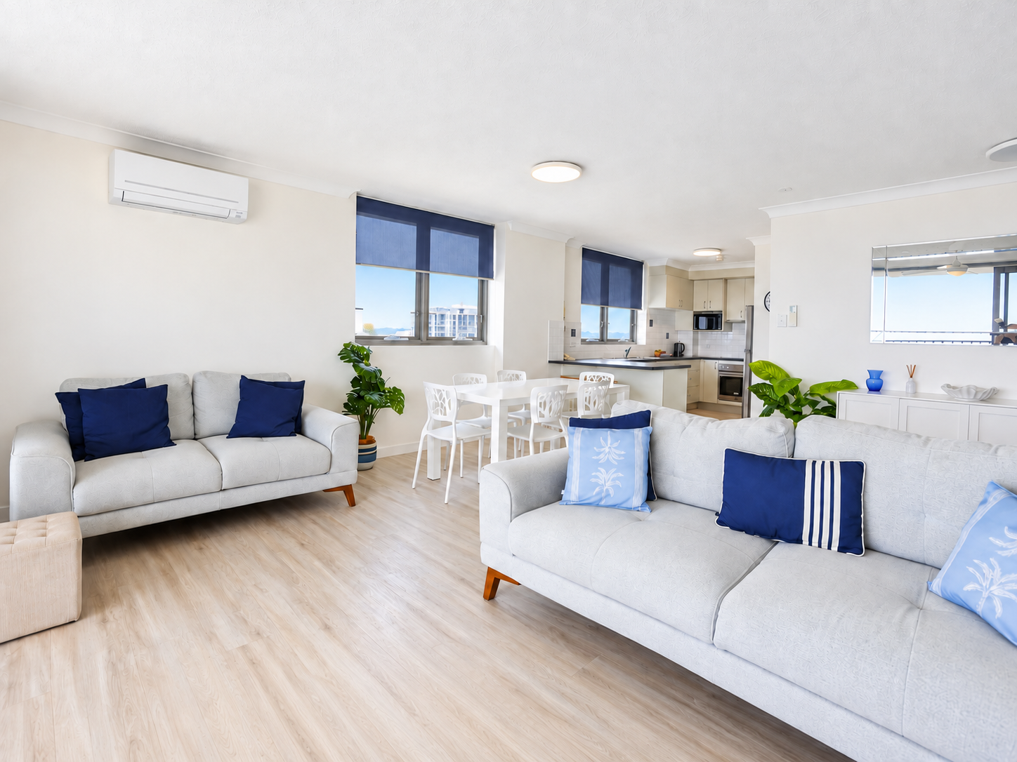 Bright living room with two light gray sofas decorated with blue and light blue cushions, a small beige ottoman, light wood flooring, and a white dining table with four white chairs near windows with blue shades. The background shows a compact kitchen with light cabinets, a microwave, a kettle, and a refrigerator, along with green potted plants and a wall-mounted air conditioner.