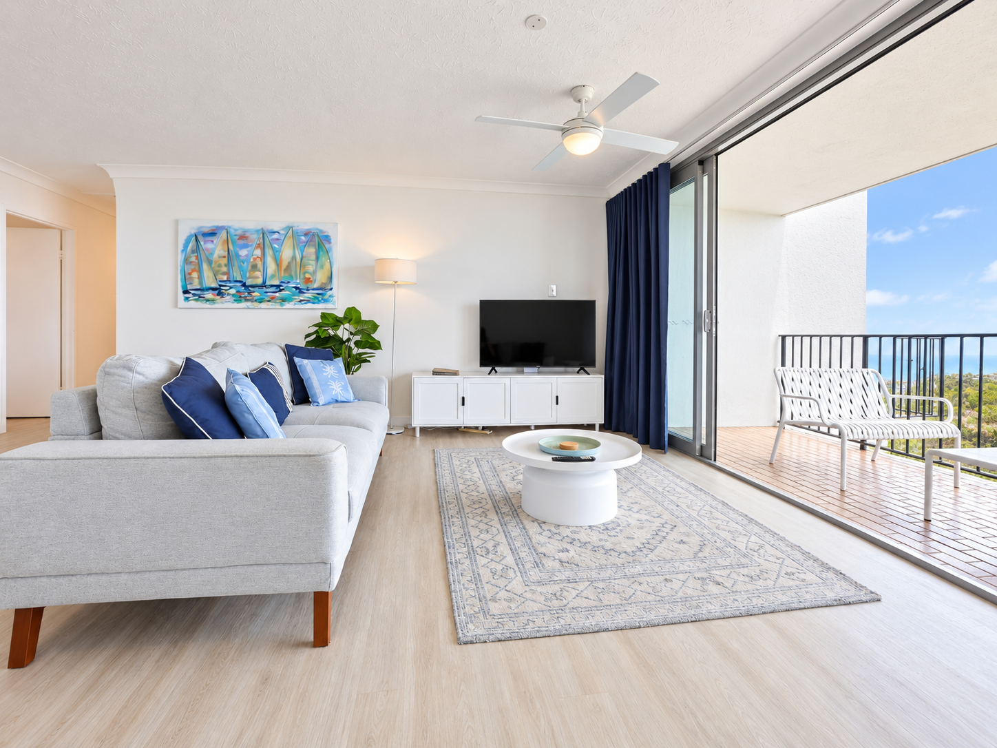 Bright living room with light wood floor, light gray sofa with blue and light blue cushions, a patterned rug, round white coffee table with decorative items, flat-screen TV on white cabinet, floor lamp, and potted plant, opening to balcony with white woven bench and view of greenery under blue sky.
