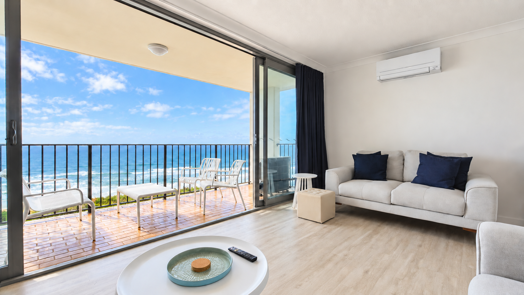 Bright living room with light wood floor, white sofa with navy blue cushions, and air conditioner on wall, opening to a balcony with four white chairs and a chaise lounge overlooking a blue ocean under a partly cloudy sky.