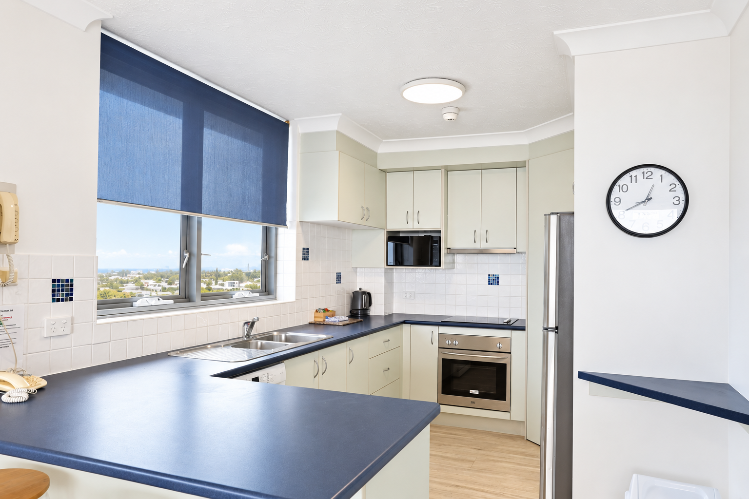 Modern kitchen with blue countertops, cream cabinets, stainless steel oven and fridge, a double sink under a window with a blue roller blind, a wall clock showing 1:10, and light wood flooring.