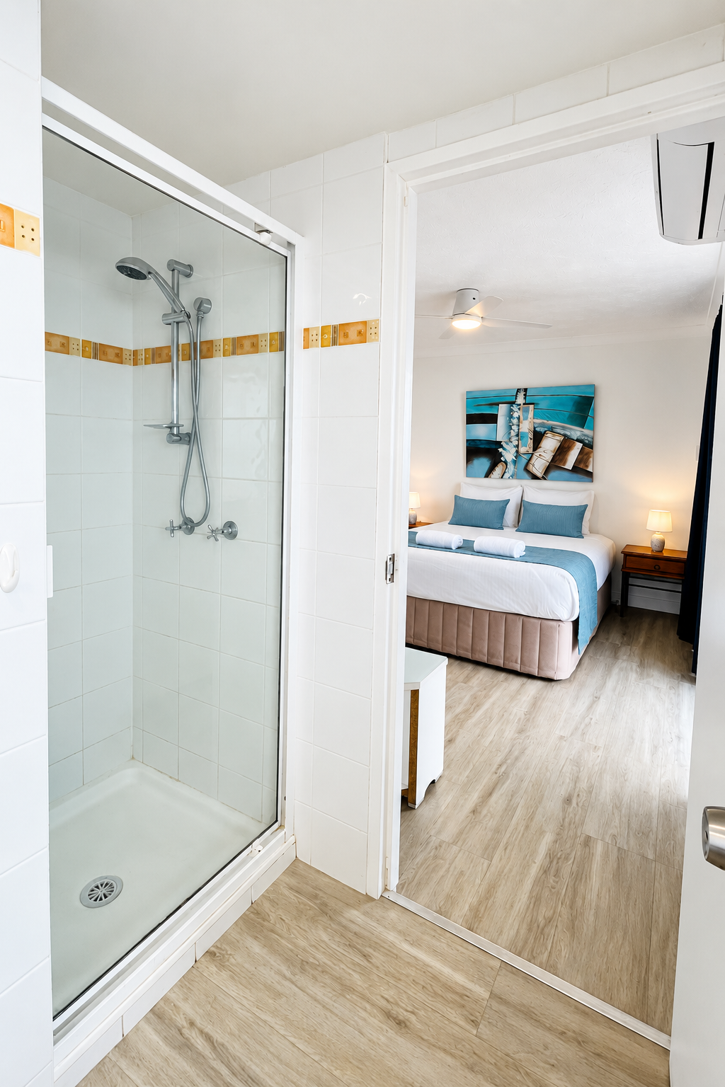 View from a bathroom featuring a tiled shower with glass door and silver showerhead, looking into a bedroom with a neatly made bed with white and blue linens, two bedside tables with lamps, a ceiling fan, and a blue abstract painting on the wall.