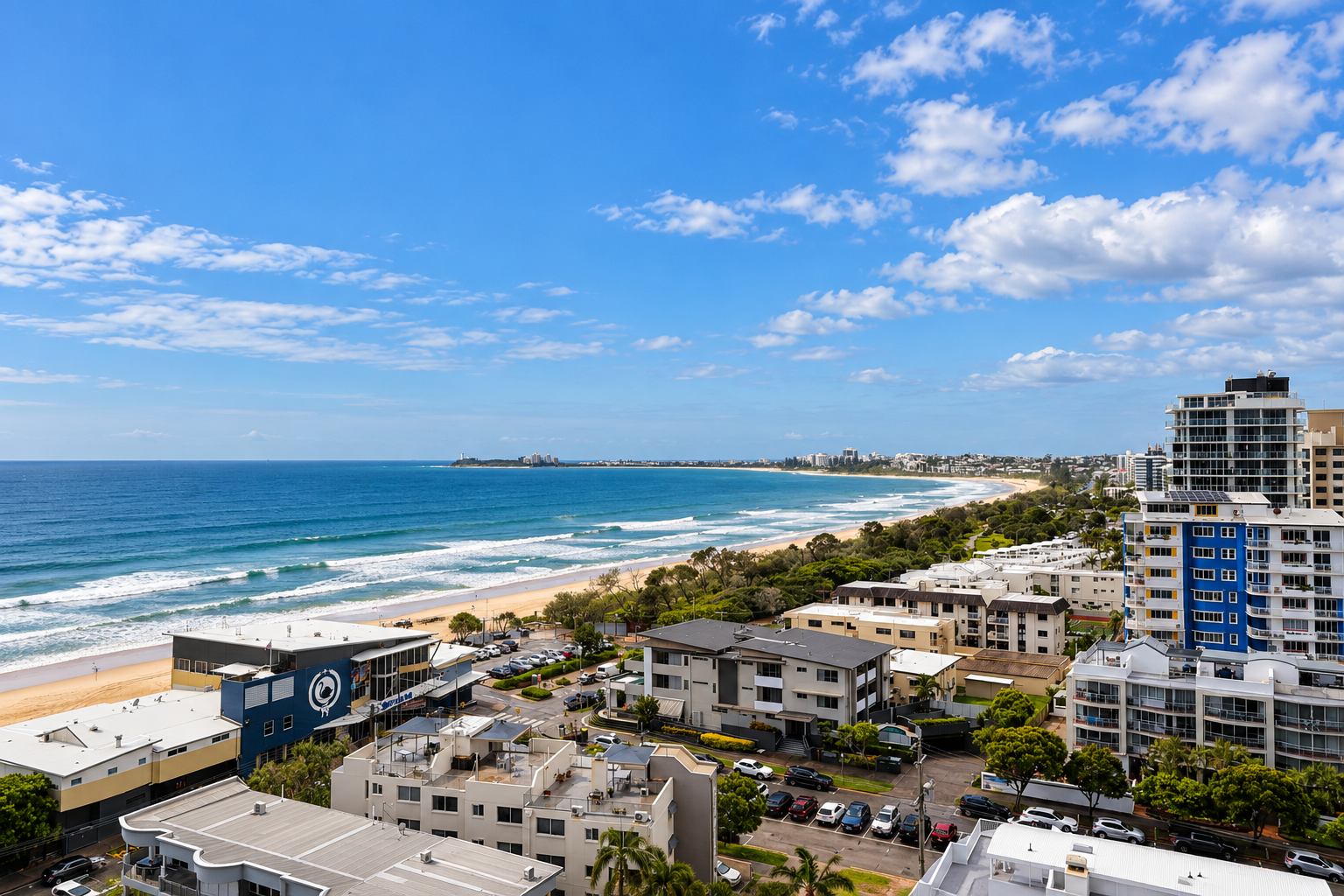 Wide coastal cityscape view with sandy beach, blue ocean waves, residential and high-rise buildings, and a partly cloudy sky.