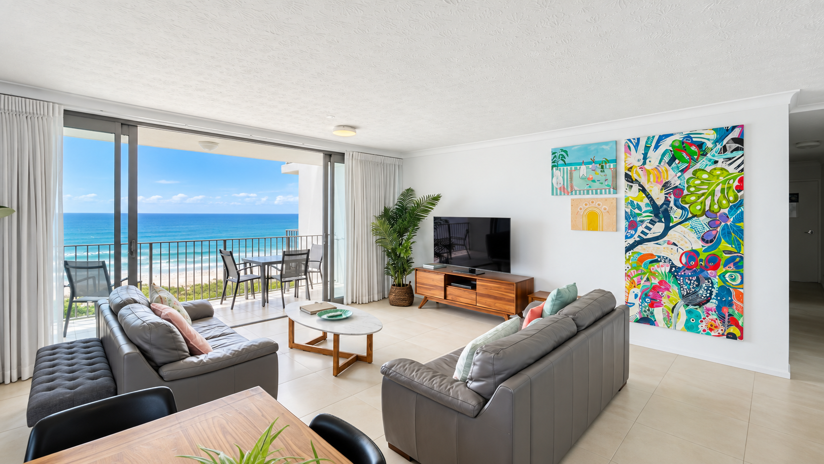 Modern living room with two gray leather sofas, a wooden coffee table, and a TV on a wooden console, featuring colorful abstract paintings on the wall and a large potted plant, with sliding glass doors opening to a balcony overlooking a blue ocean and sandy beach.