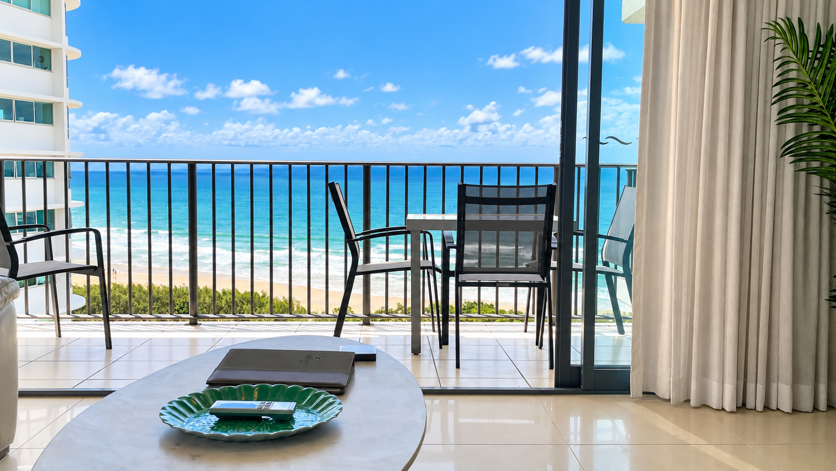 View from a tiled living room with sliding glass doors opening to a balcony overlooking a sandy beach and blue ocean under a clear sky with scattered clouds; the balcony has a white table and four black chairs, and a green decorative plate with remote controls sits on a white coffee table inside.