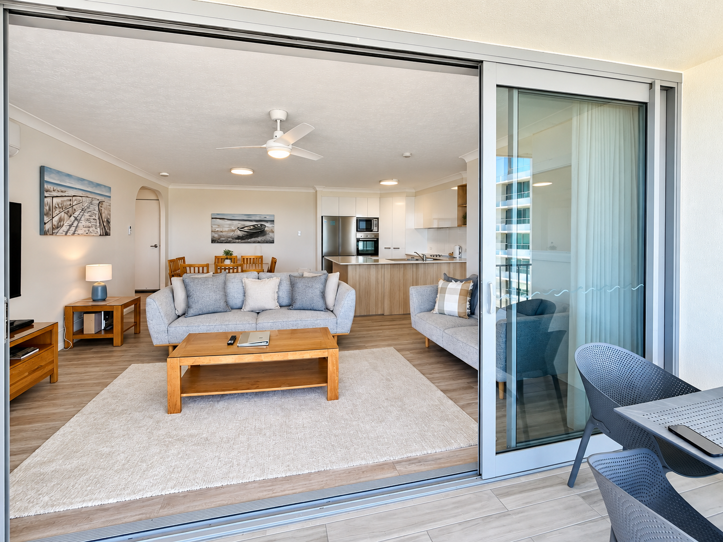 Open-plan living space viewed from balcony, featuring two light gray sofas with cushions, wooden coffee table, dining table with chairs, modern kitchen with white cabinets and wood accents, ceiling fan, and wall art.
