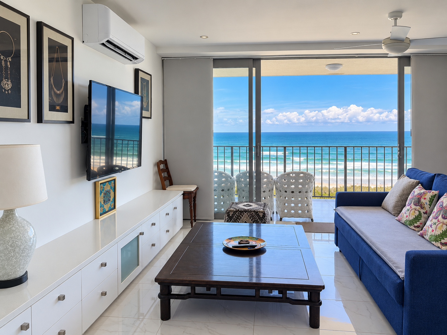 Modern living room with a blue sofa adorned with colorful pillows, a dark wooden coffee table with a decorative plate, a white entertainment unit with framed art, and large sliding glass doors opening to a balcony overlooking an ocean view with blue sky and waves.