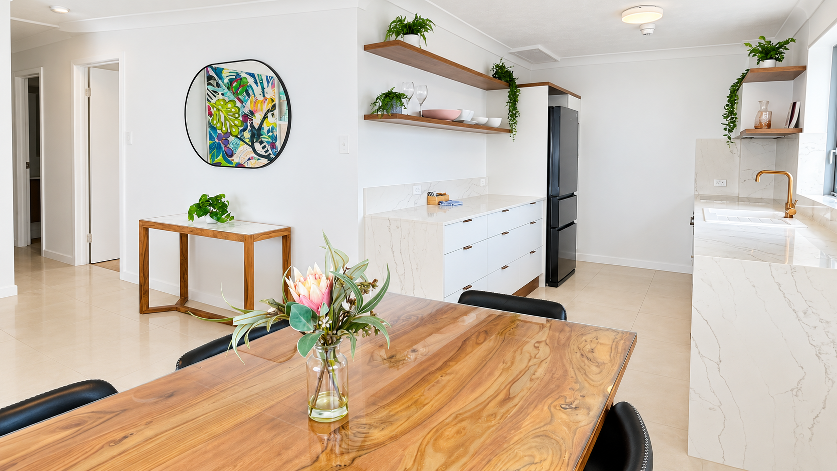 Modern kitchen and dining area with a polished wooden dining table with a glass vase of flowers, white marble countertops, gold faucet, black fridge, wooden floating shelves with plants and dishes, and a small wooden side table under a round colorful mirror on white walls.