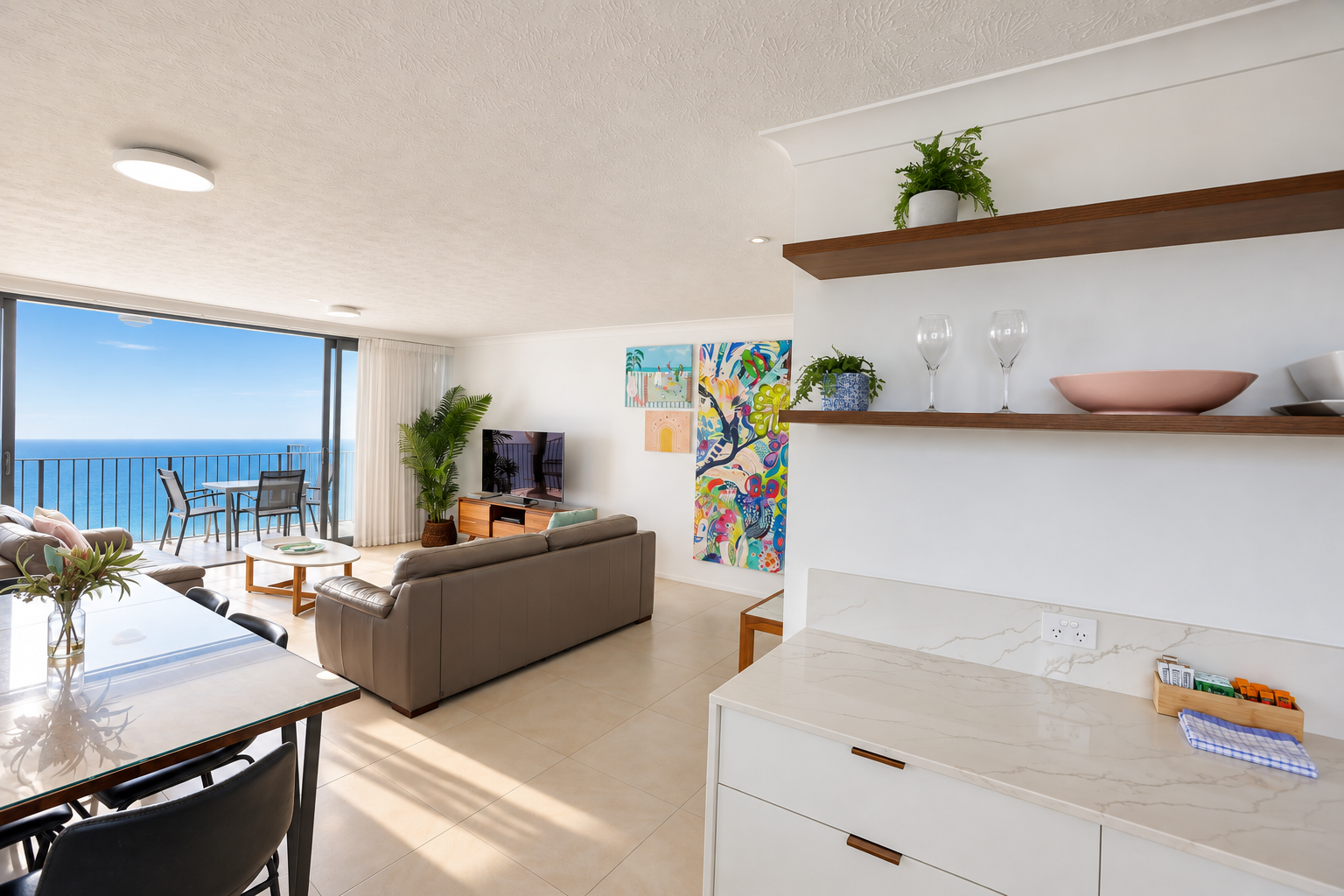 Modern open-plan living area with beige tiled floor featuring a glass-top dining table with black chairs on the left and two brown sofas facing a TV on a wooden stand against the far wall. Sliding glass doors open to a balcony with outdoor seating and a vast ocean view under a clear blue sky. On the right, white kitchen countertops and cabinetry with wooden floating shelves hold plants, two wine glasses, and bowls. Bright natural light fills the space.