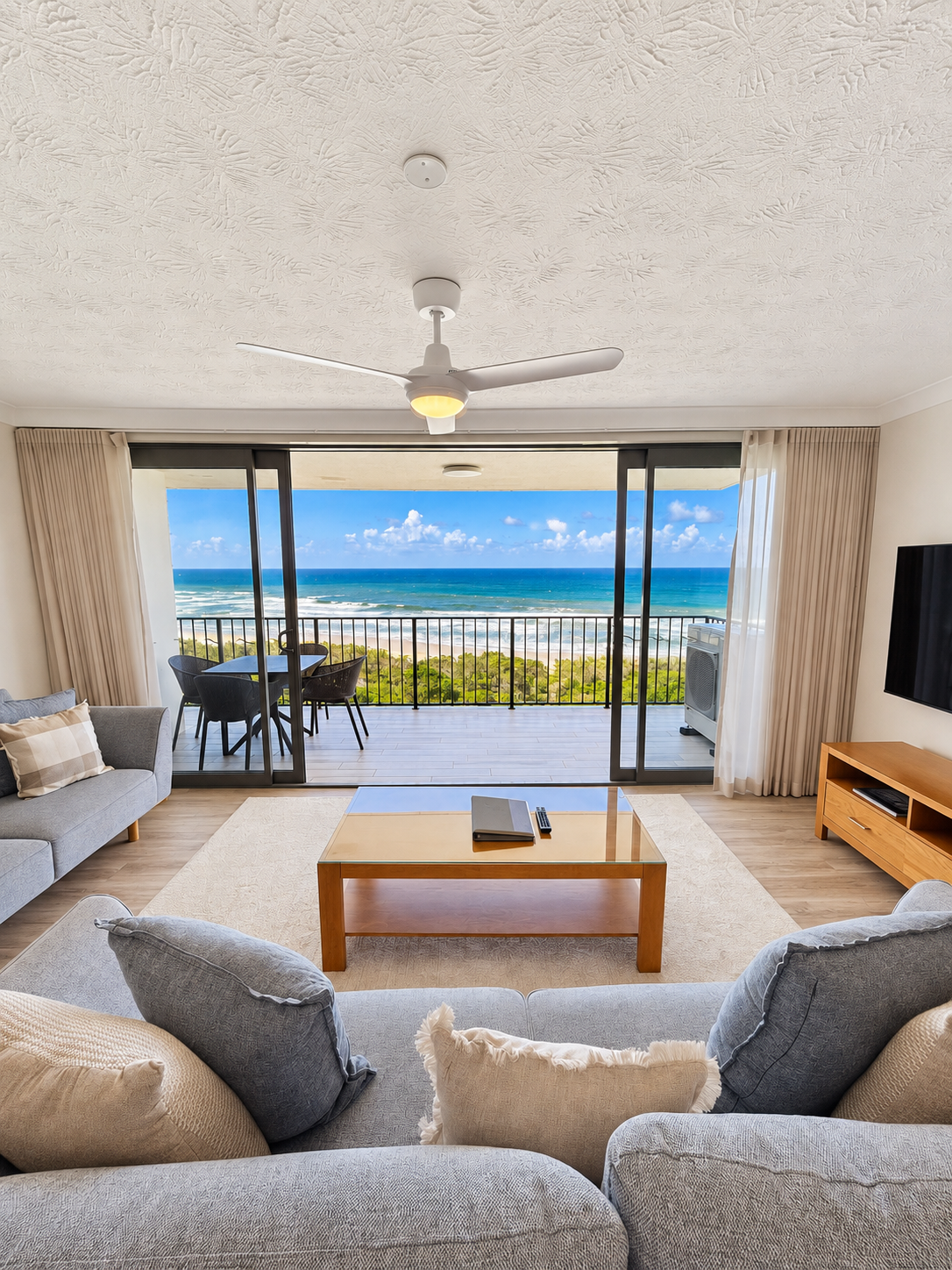 Bright living room with gray sofas and cushions facing a glass coffee table, sliding doors opening to a balcony with outdoor seating overlooking the ocean under a blue sky with clouds.