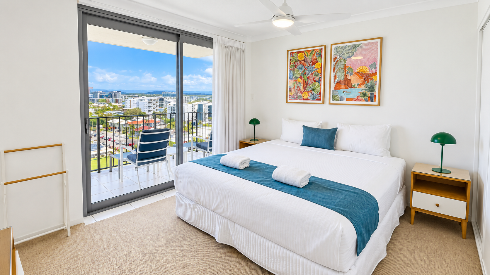 Bright master bedroom with a large bed dressed in white linens and a teal runner, two rolled white towels on the bed, a teal pillow, two wooden nightstands each with a green lamp, colorful framed artwork above the bed, a ceiling fan, and a sliding glass door leading to a balcony with two blue cushioned chairs overlooking a cityscape under a blue sky.