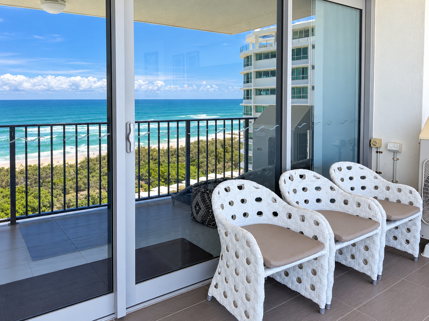Balcony with three white woven chairs having beige cushions, sliding glass doors, a railing, and a view of the beach, ocean, and blue sky with clouds.