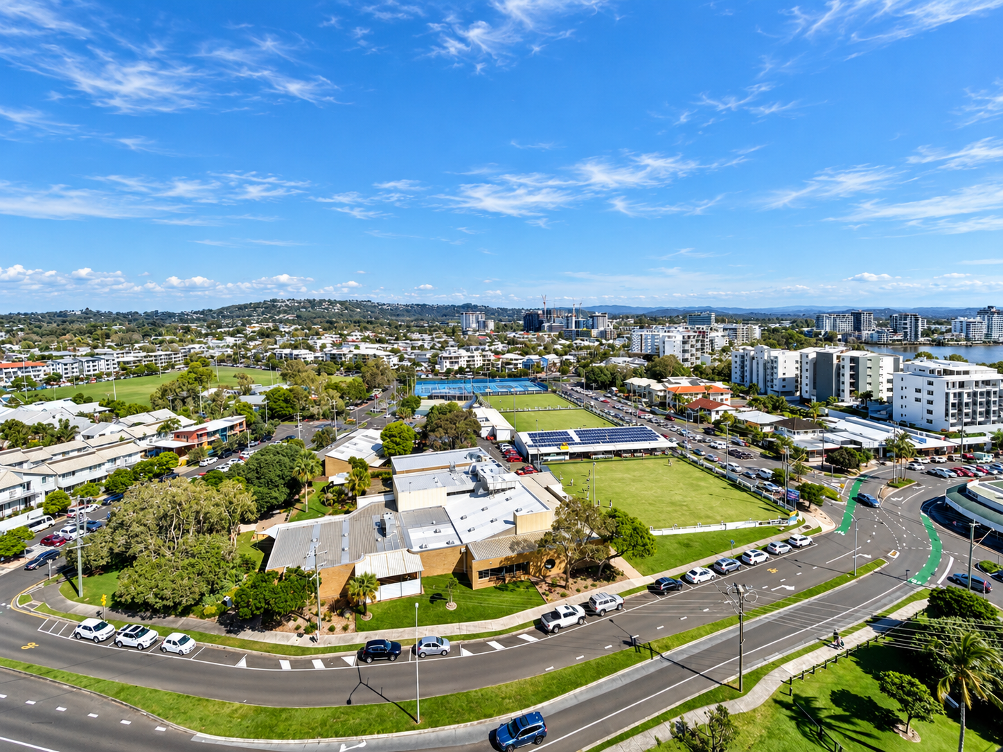 Aerial view of a suburban area with residential buildings, green sports fields, trees, and a partly cloudy sky, featuring roads with parked and moving cars surrounding the neighborhood.