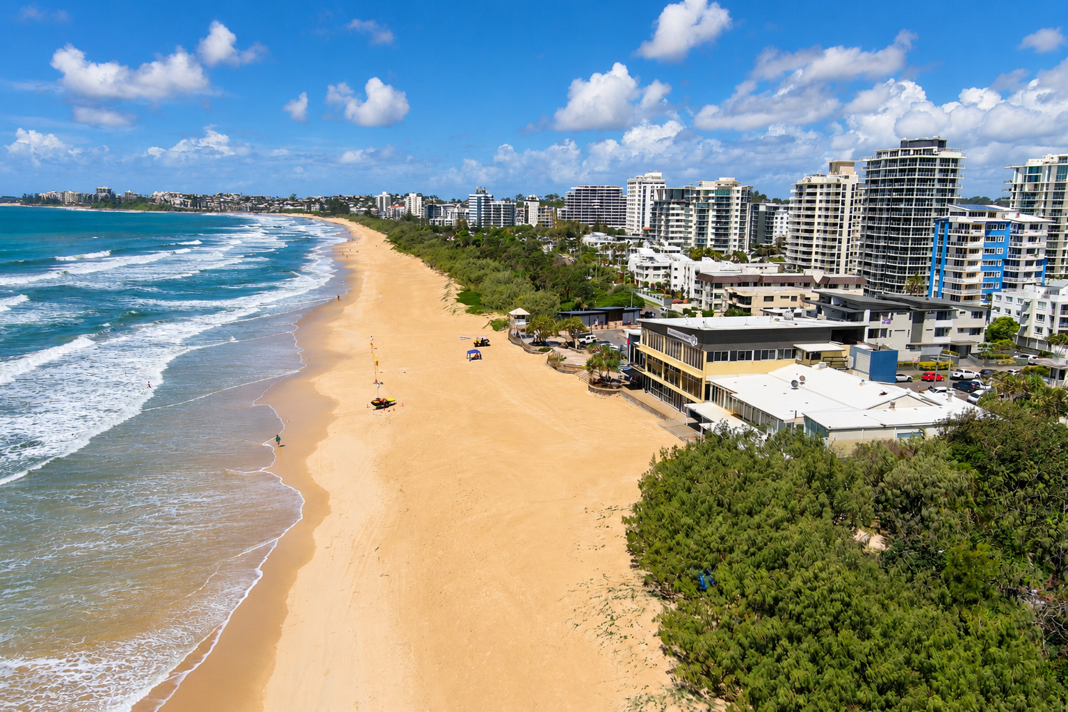 Aerial view of a wide sandy beach with gentle waves along the shore, bordered by green trees and a row of modern high-rise buildings under a blue sky with scattered clouds.