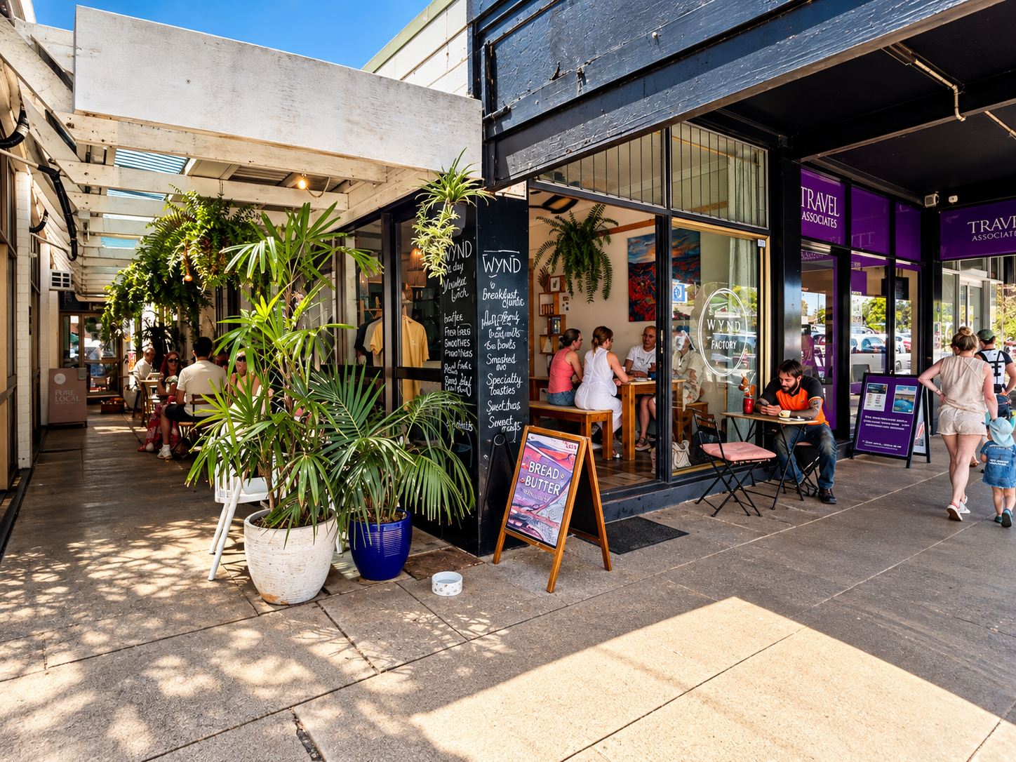 Outdoor view of a bustling café called Wynd Factory with people sitting both inside and outside, surrounded by potted plants and hanging greenery, with menu boards near the entrance and purple signage for nearby Travel Associates offices.