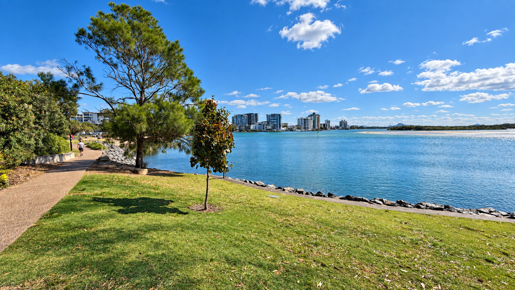 Riverfront park with a paved walking path, green grass, scattered trees, and a large river reflecting a blue sky with white clouds, with distant modern buildings across the water.