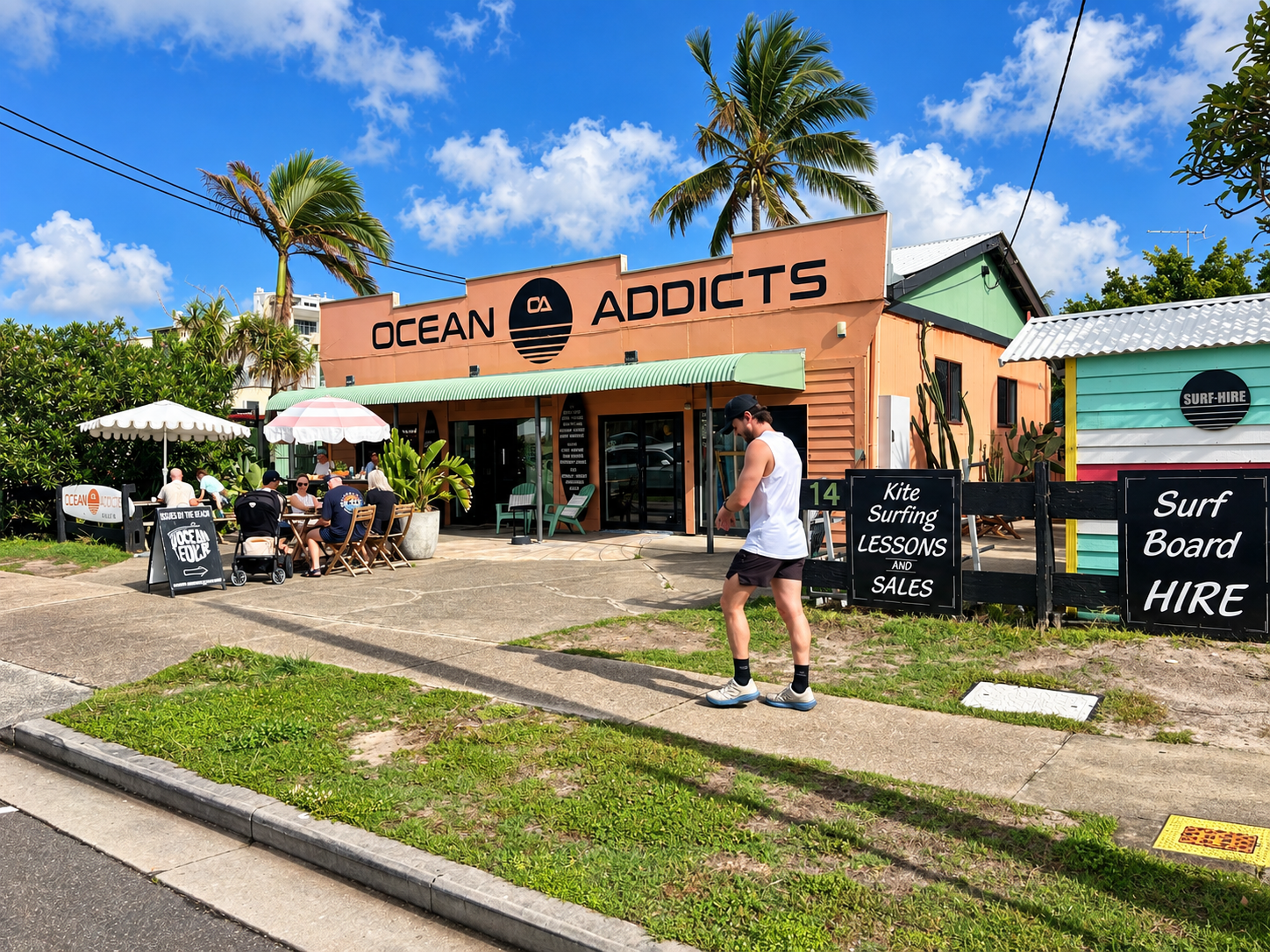 People sitting under umbrellas at wooden tables outside a building with a sign reading Ocean Addicts. A man in athletic wear walks on the sidewalk past signs advertising kite surfing lessons and surfboard hire. The scene includes palm trees, blue sky with clouds, and a colorful surf-hire shack.