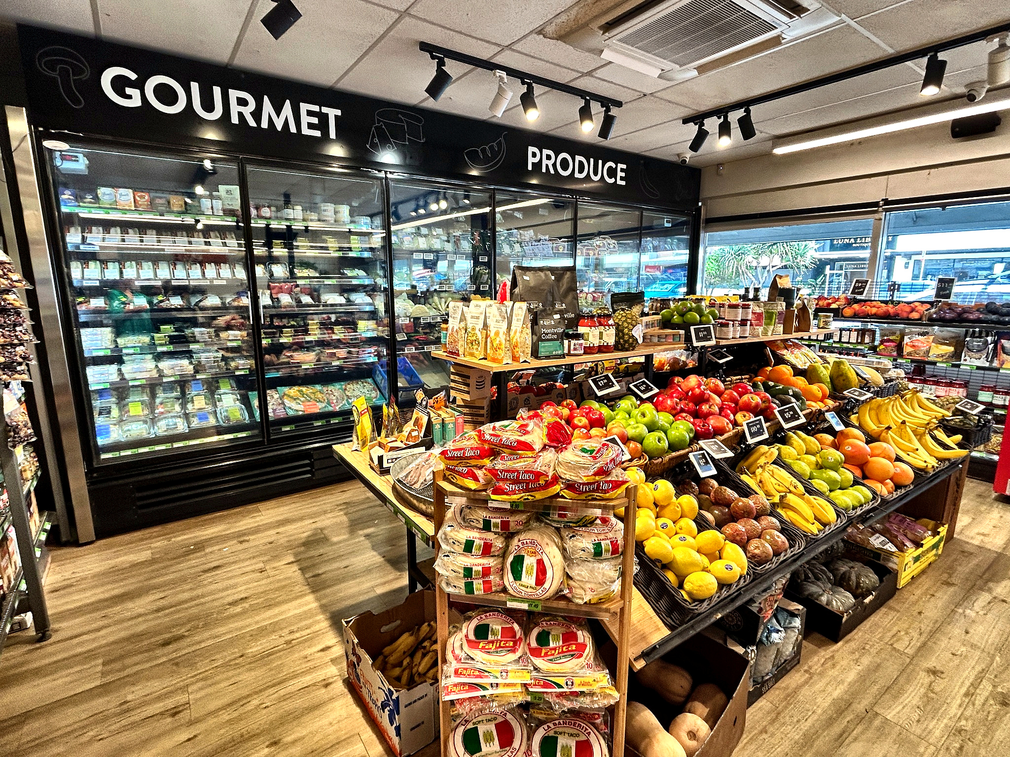 Interior of a gourmet produce grocery with a central display of fresh fruits including apples, bananas, oranges, and avocados, surrounded by packaged goods and refrigerated sections.