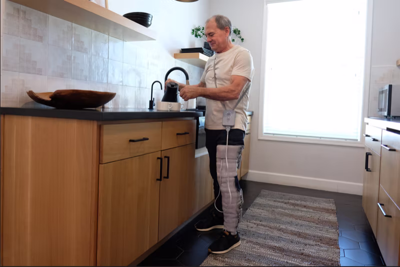 Man standing in his kitchen pouring some coffee with the Dayspring lower delivering treatment to his leg 
