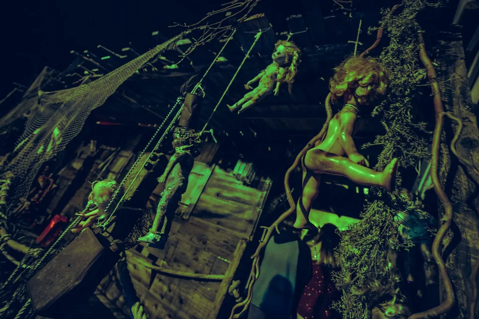 Hanging dolls and tangled ropes fill a wooden haunt set under green light.