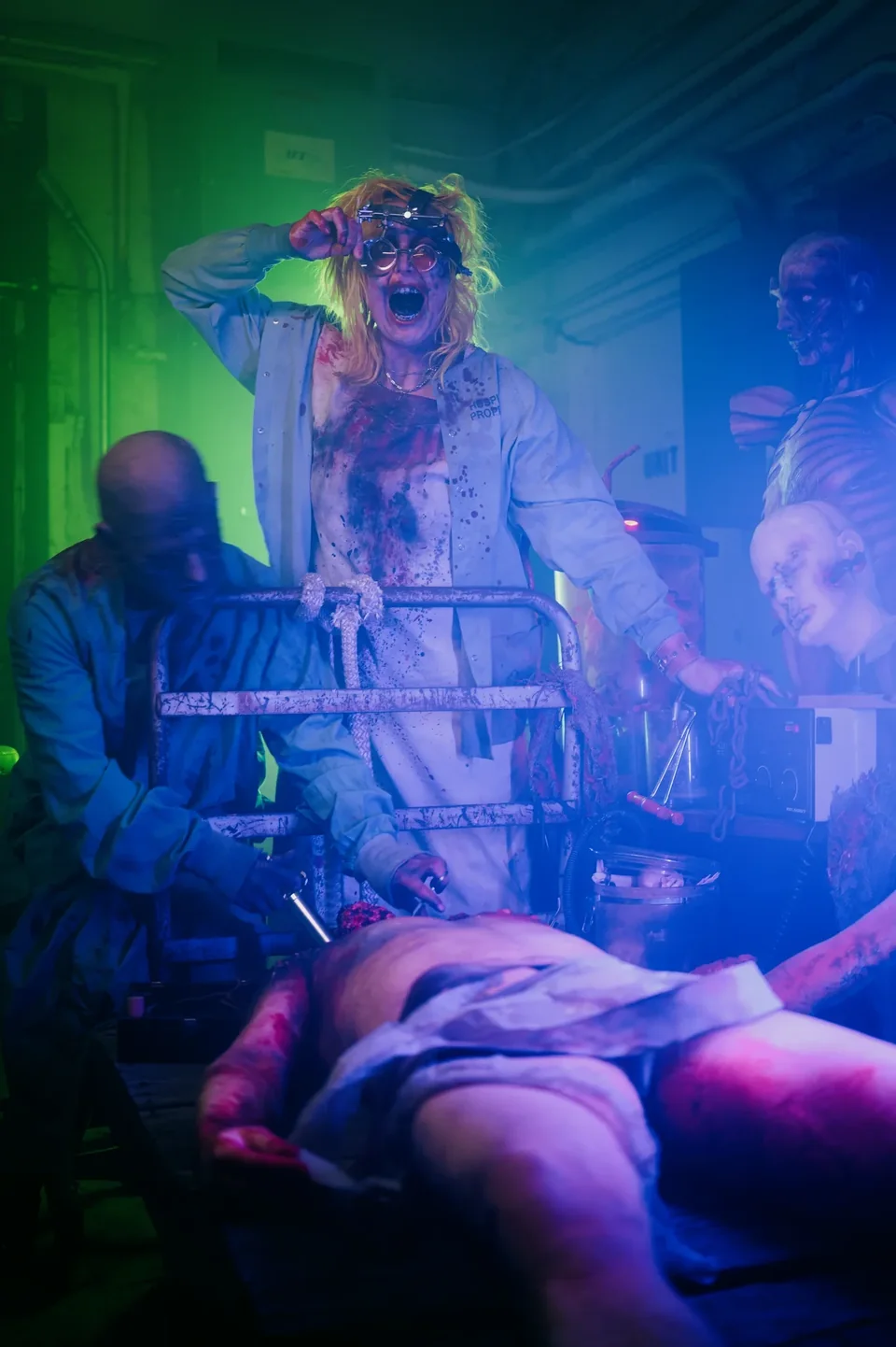 A goggled laboratory performer and assistants in green and blue light.