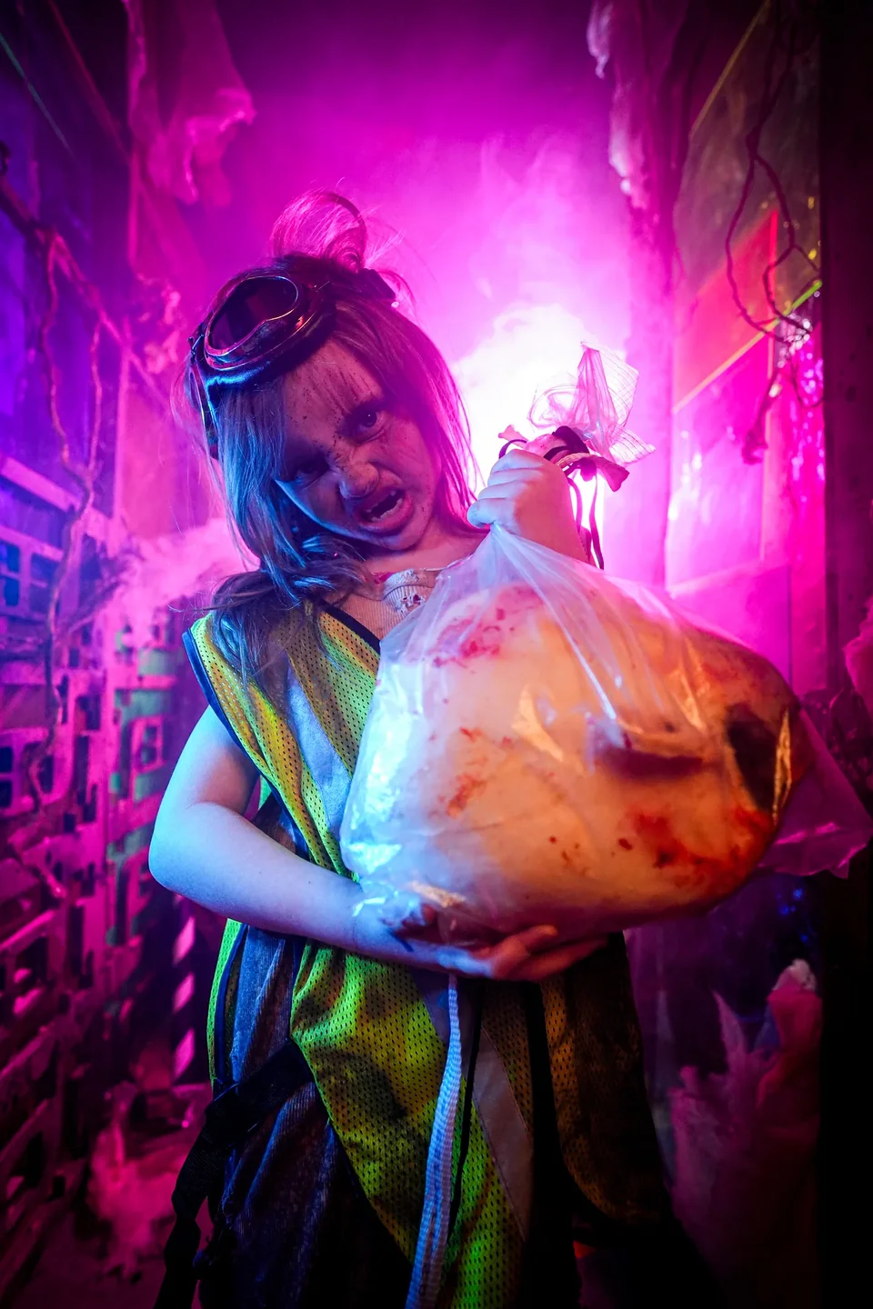 A goggled performer holds a bagged theatrical prop in a passage glowing magenta and blue.