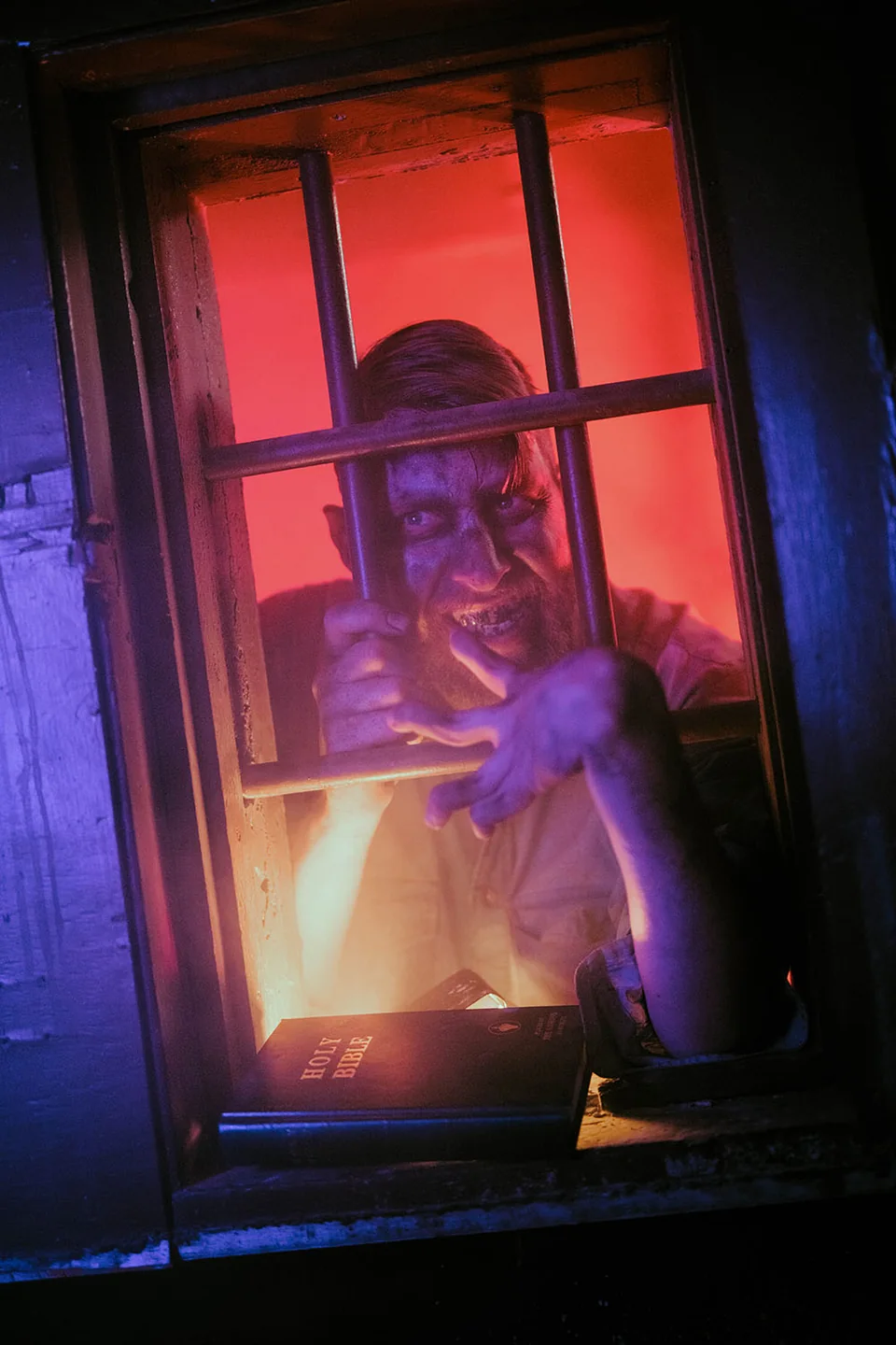A bearded prisoner peers through a barred window lit orange and violet.