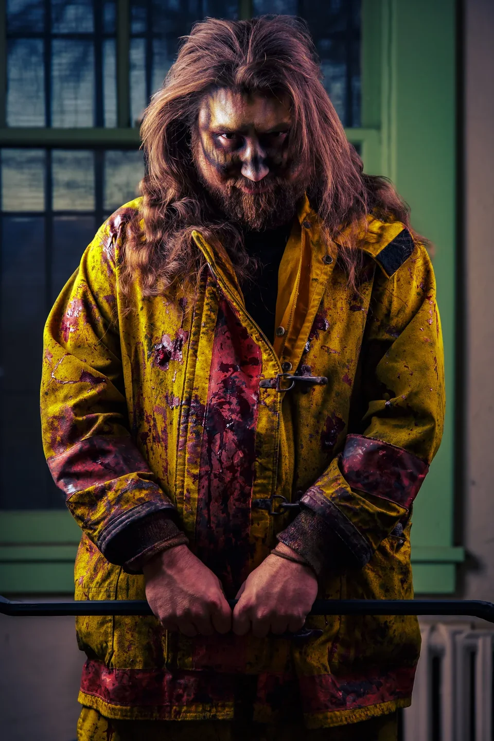 A long-haired performer in a yellow firefighter coat stares out from in front of green prison bars.