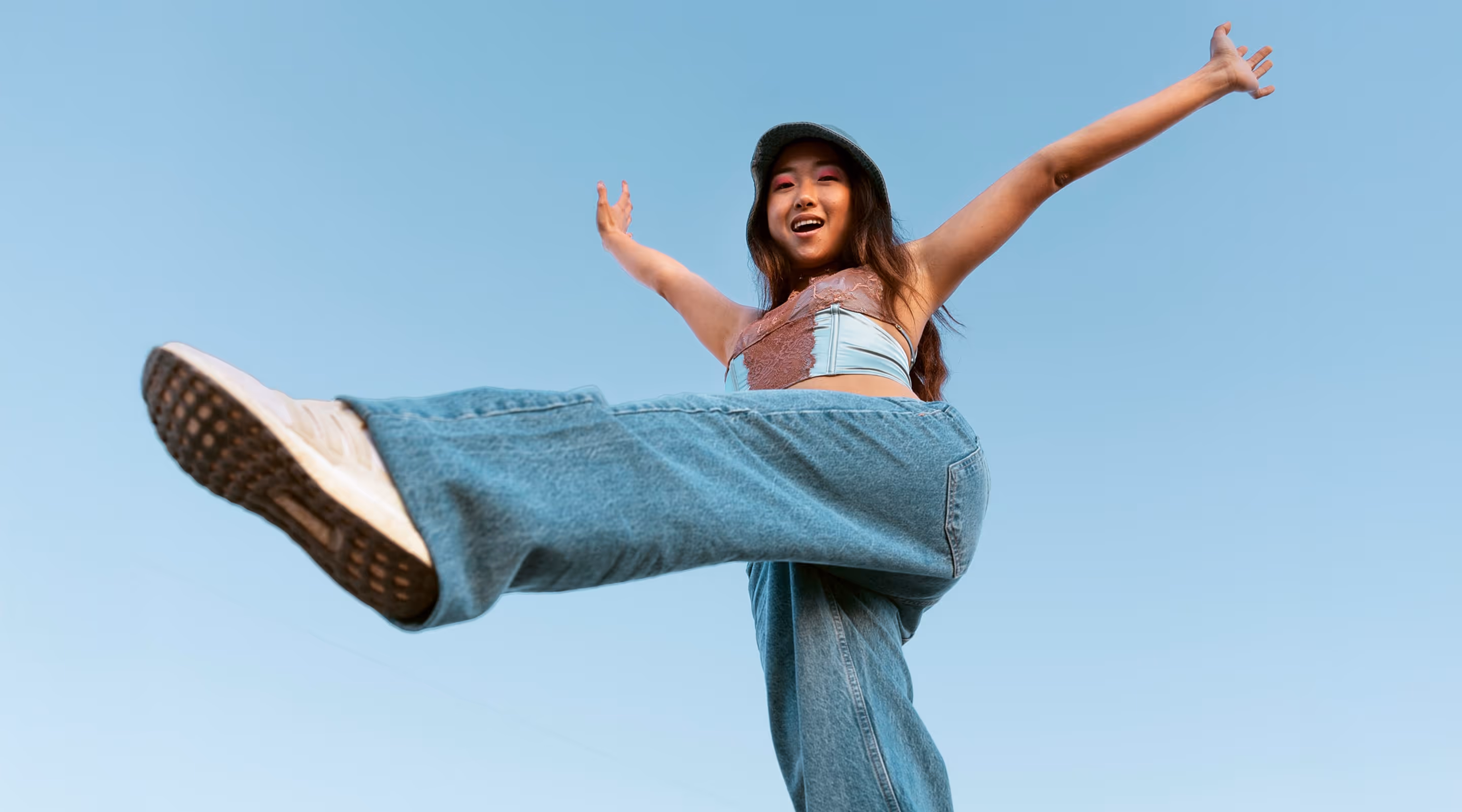 Young woman in jeans and a top joyfully kicking a leg forward against a clear blue sky.