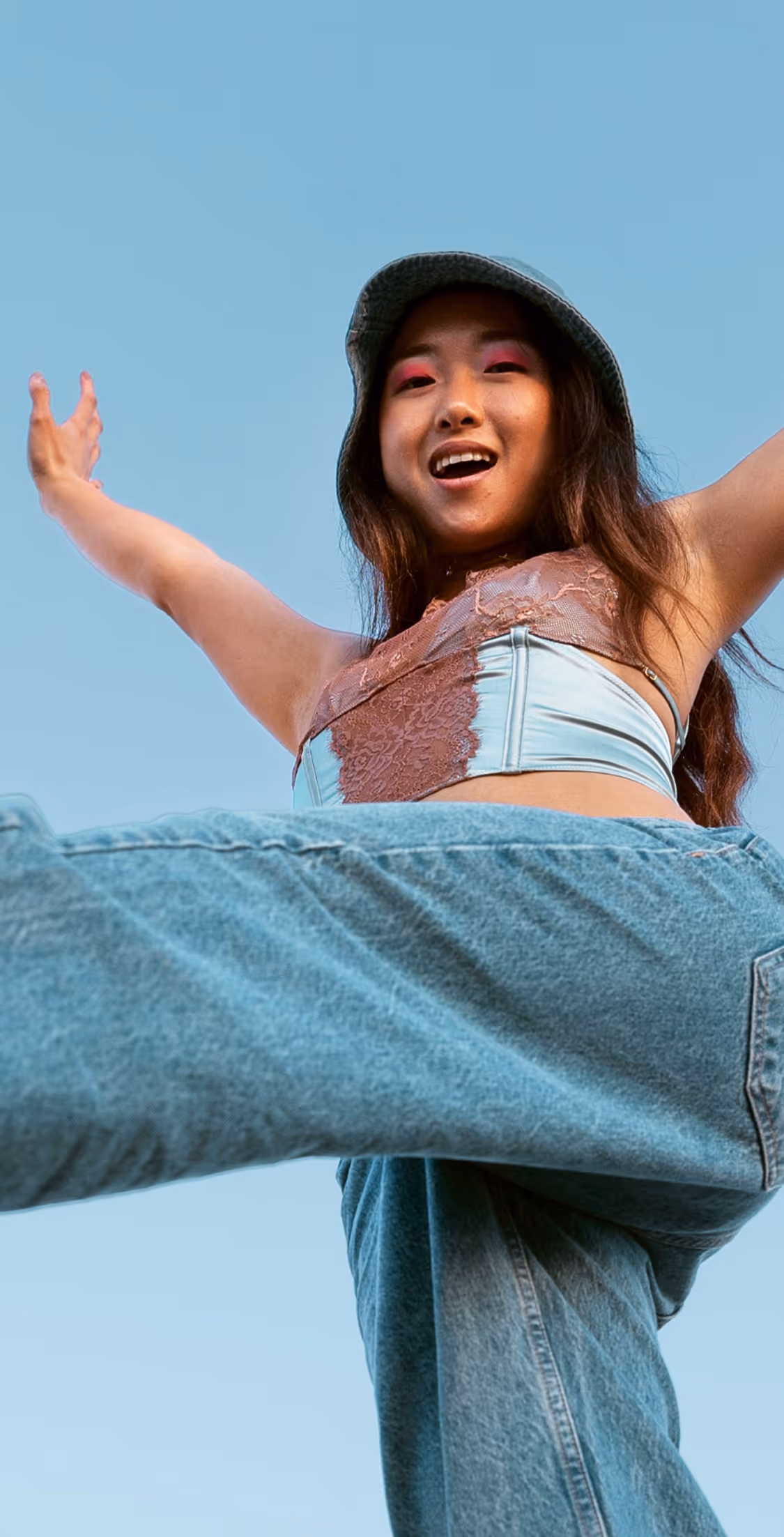 Low-angle view of a woman wearing a bucket hat and colorful top with arms outstretched against a clear blue sky.