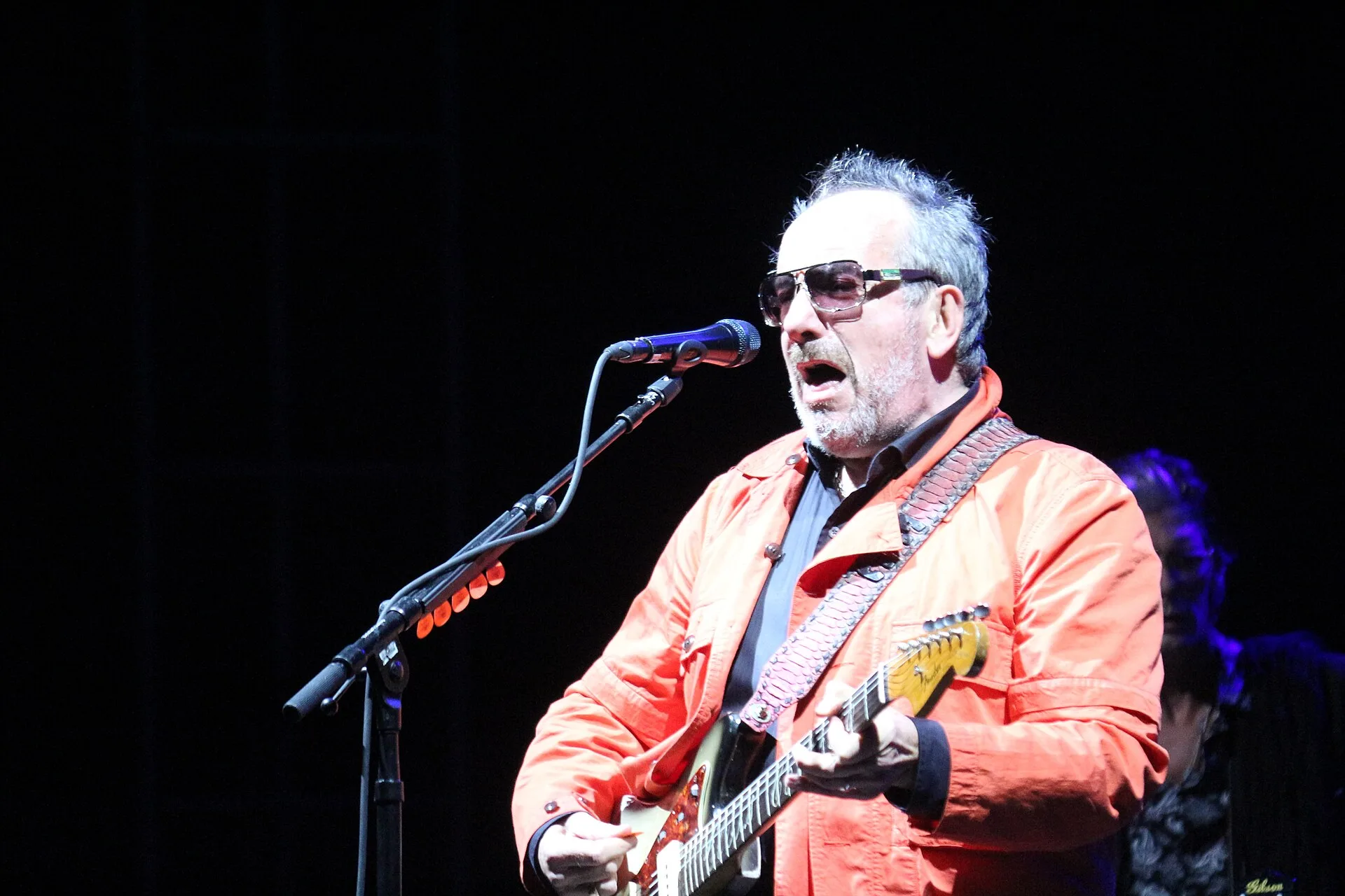 Elvis Costello in an orange jacket singing and playing an electric guitar
