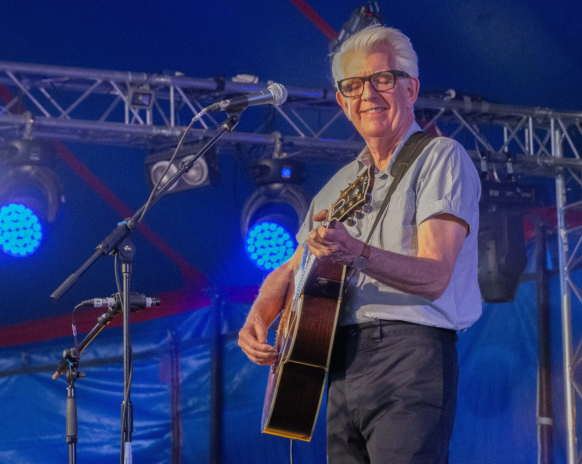 Nick Lowe singing and playing an acoustic guitar on a stage under blue lights
