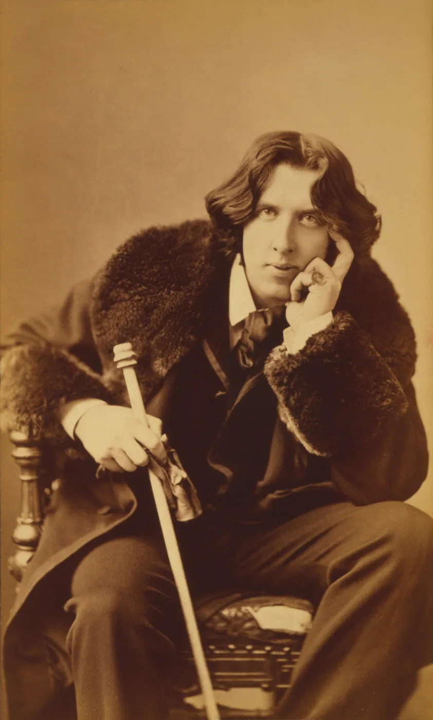 Oscar Wilde with long hair seated in a dark jacket, holding a cane