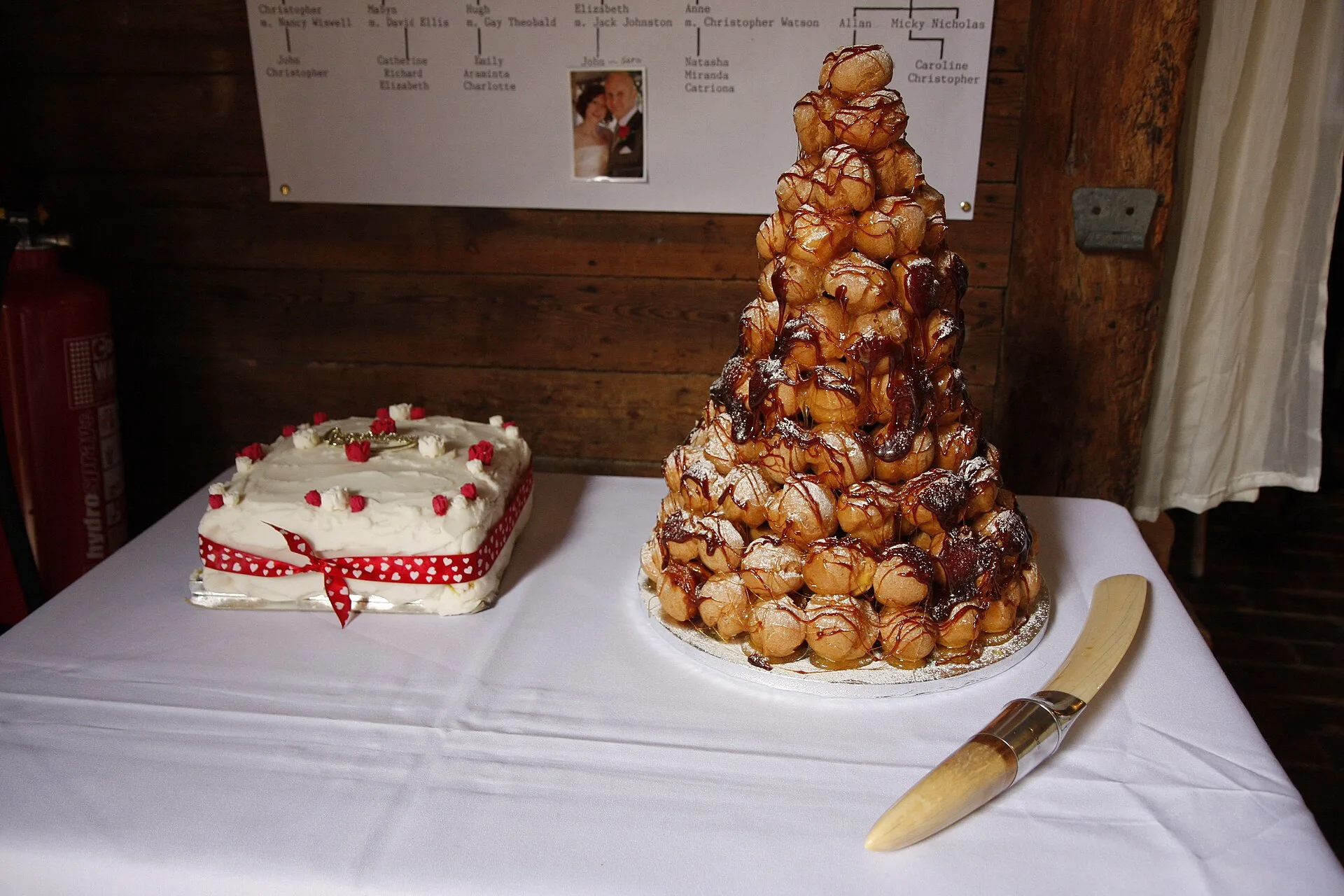 Tall cone of choux buns beside a decorated cake on a white tablecloth