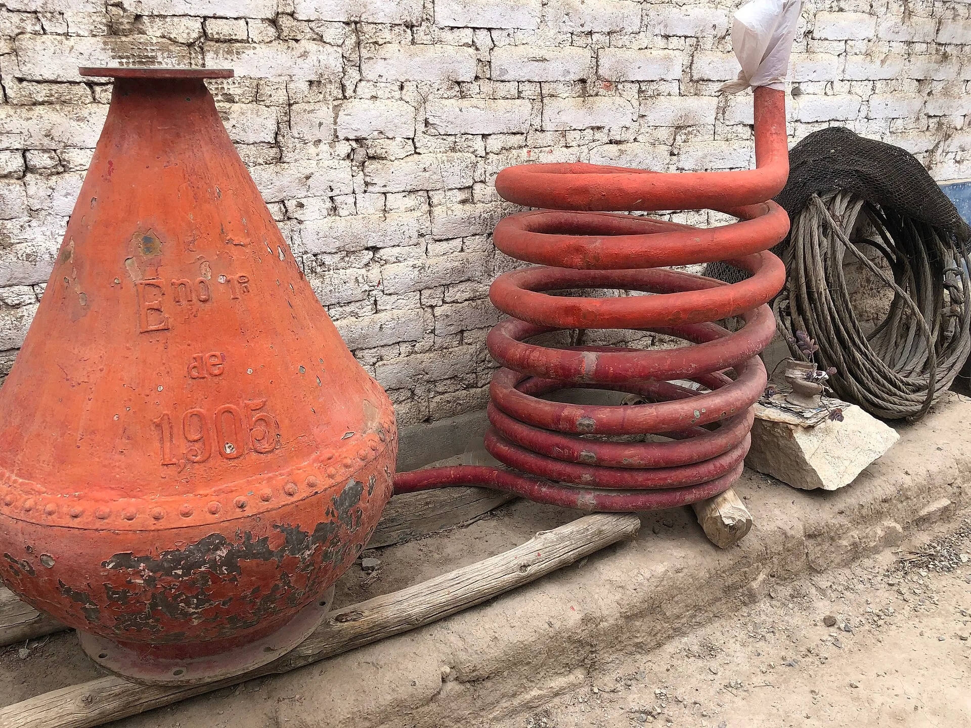 Red-painted conical still beside a coiled metal condenser against a brick wall