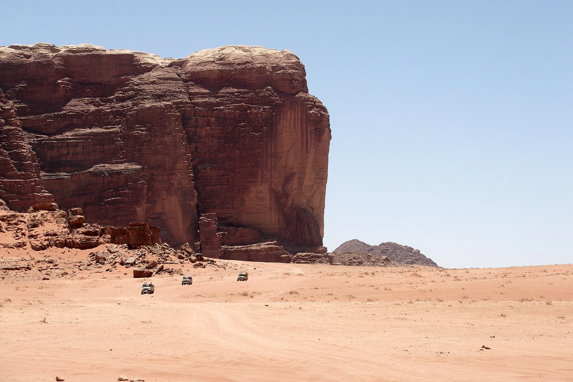 Sheer sandstone cliff rising from flat sand with small vehicles at its base