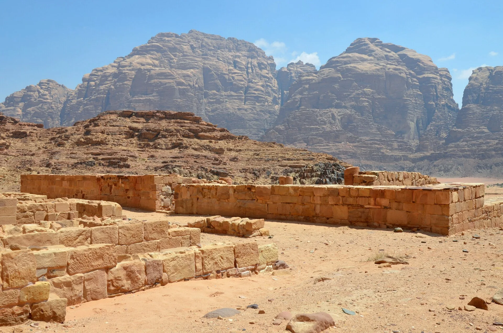 Low stone wall foundations in sand below a towering rock massif