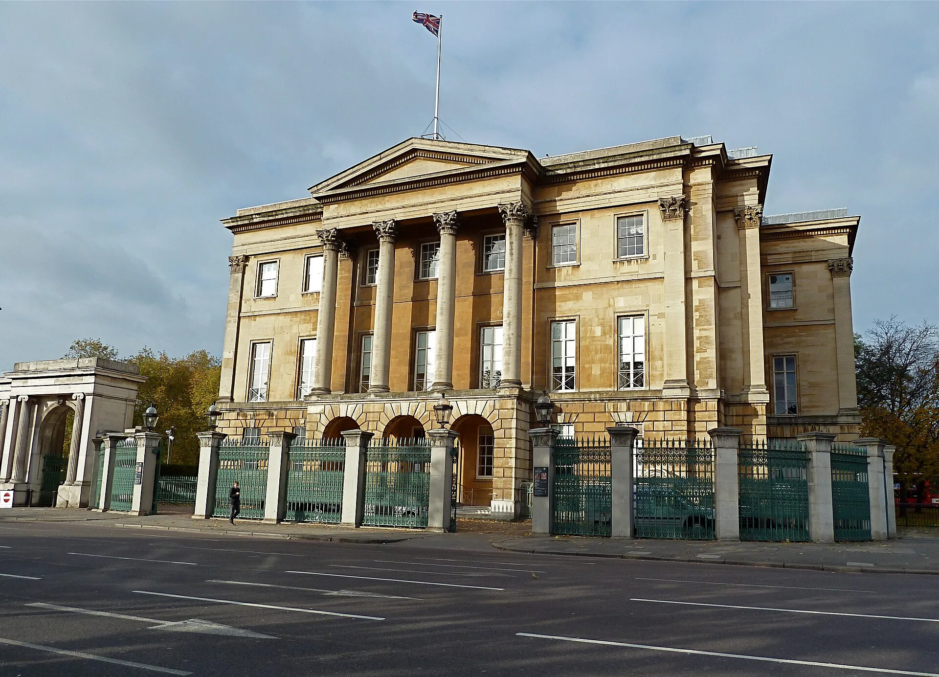 Classical stone mansion with a columned portico beside railings and a road