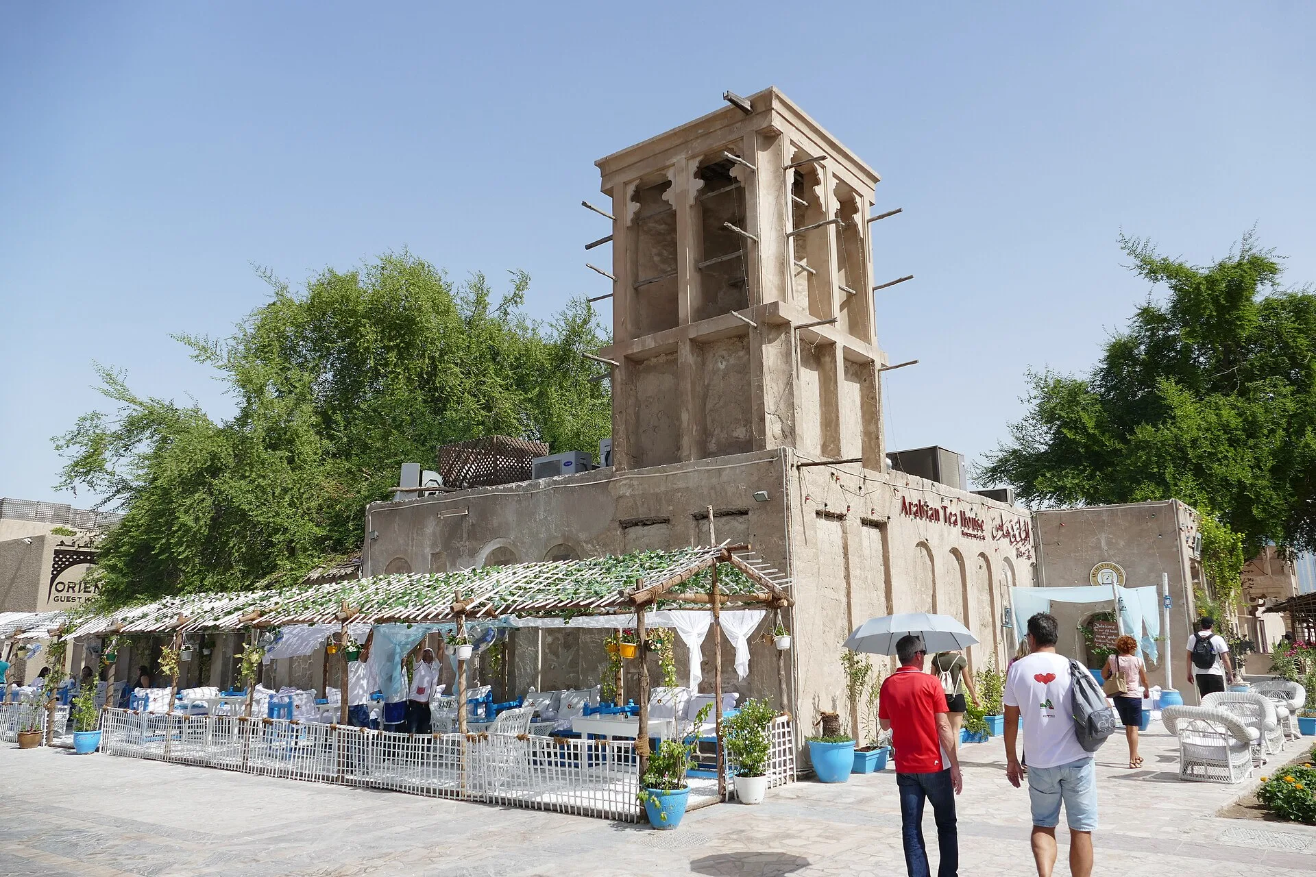 Sand-coloured house with a tall wind tower above a shaded courtyard café
