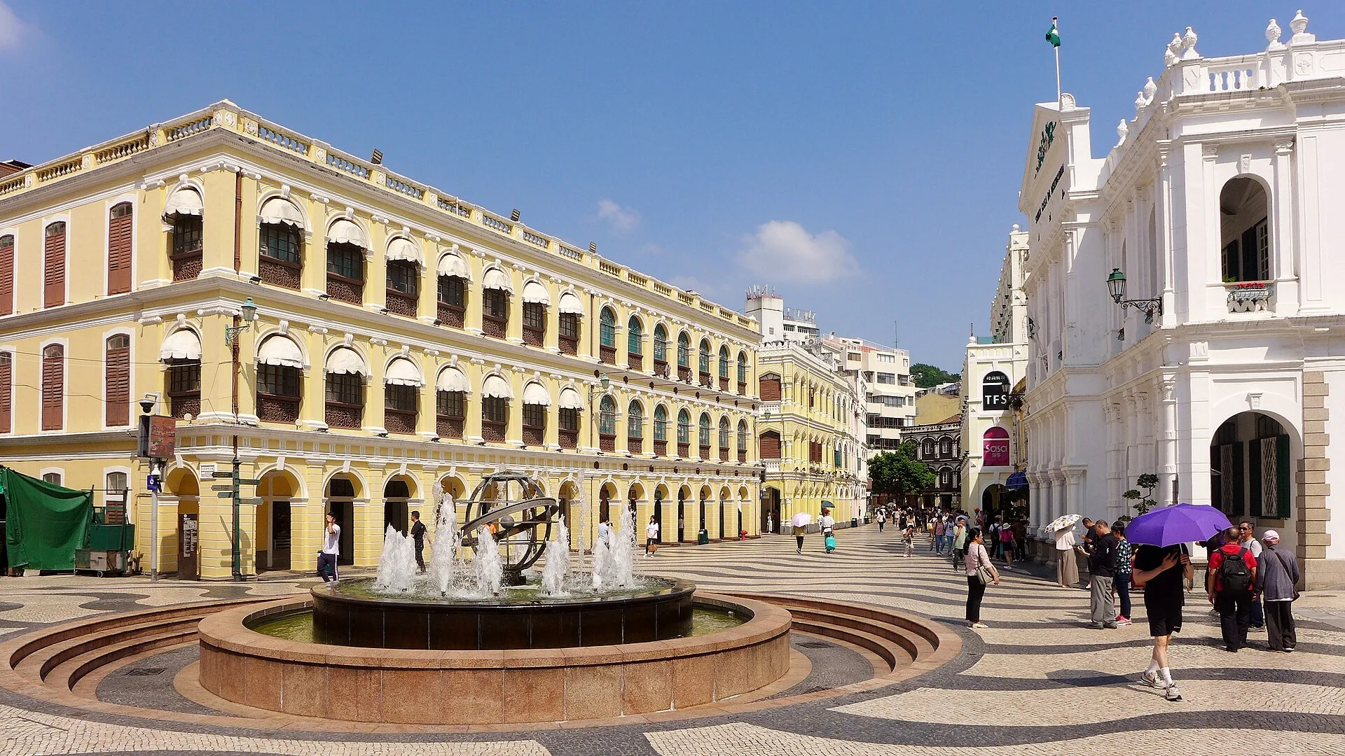 Fountain in a square with wave-patterned paving and arcaded yellow buildings