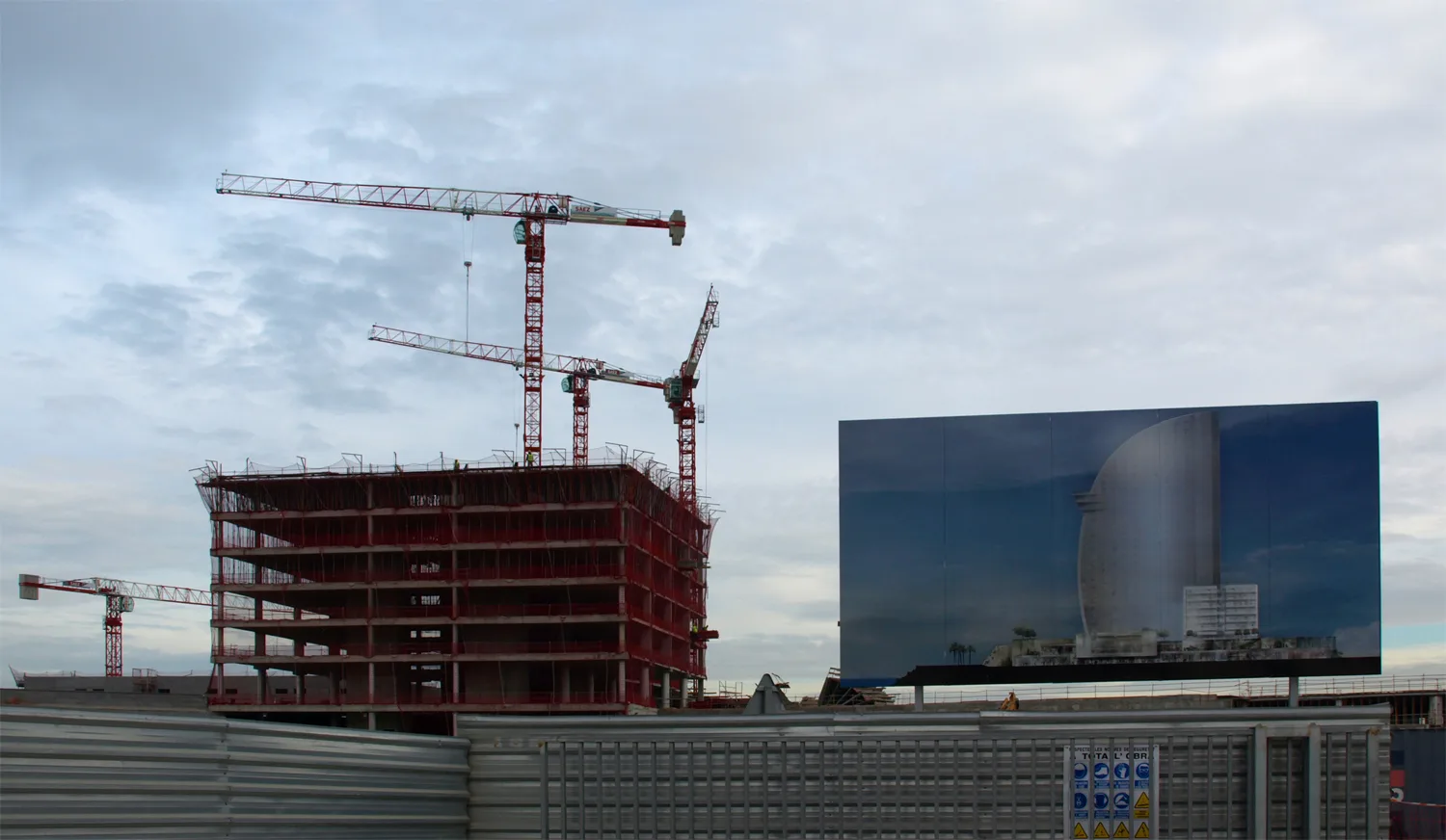Concrete building frame with tower cranes beside a large reflective hoarding