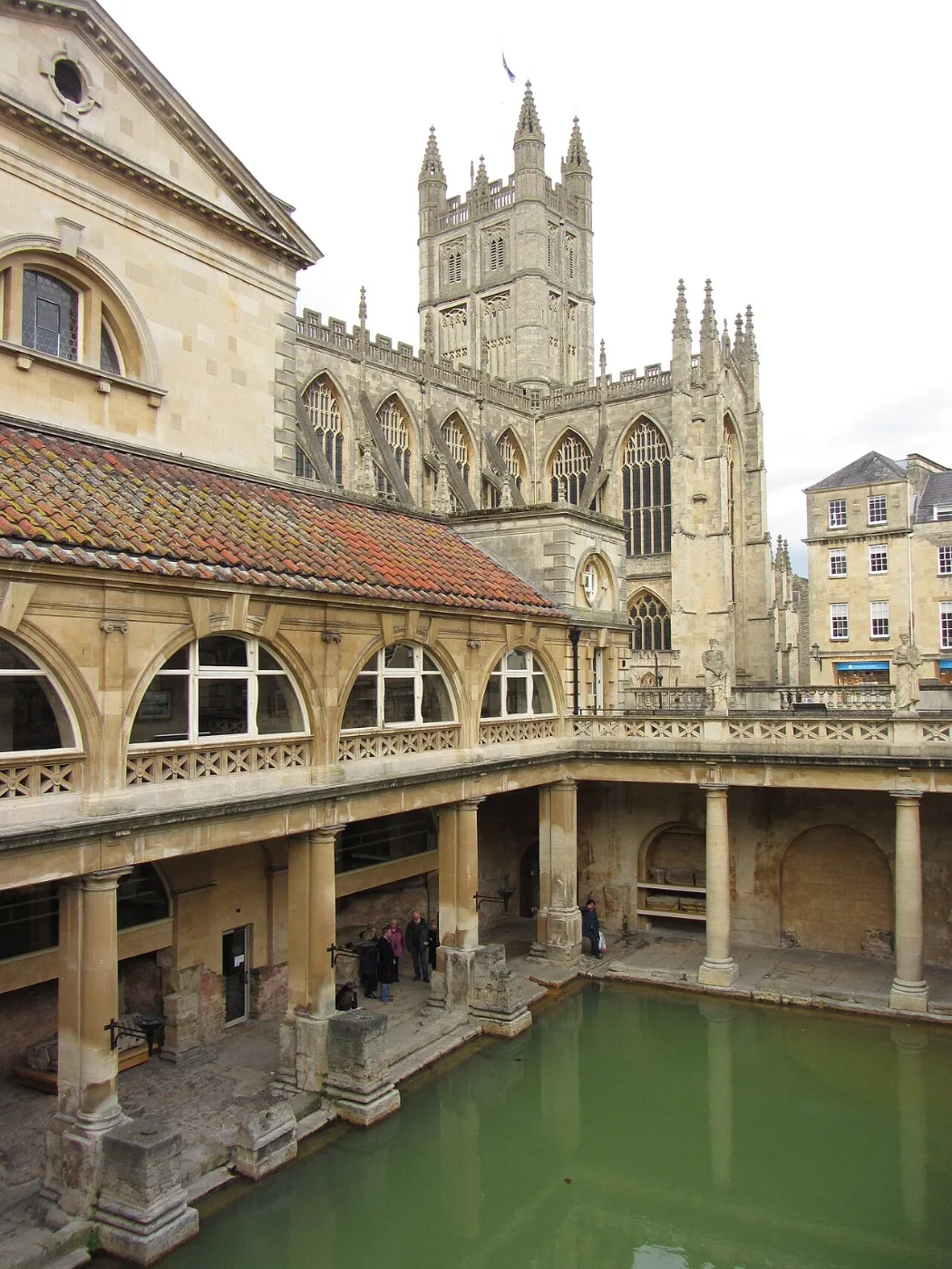 Green water of a Roman bath beneath a colonnaded terrace and abbey tower