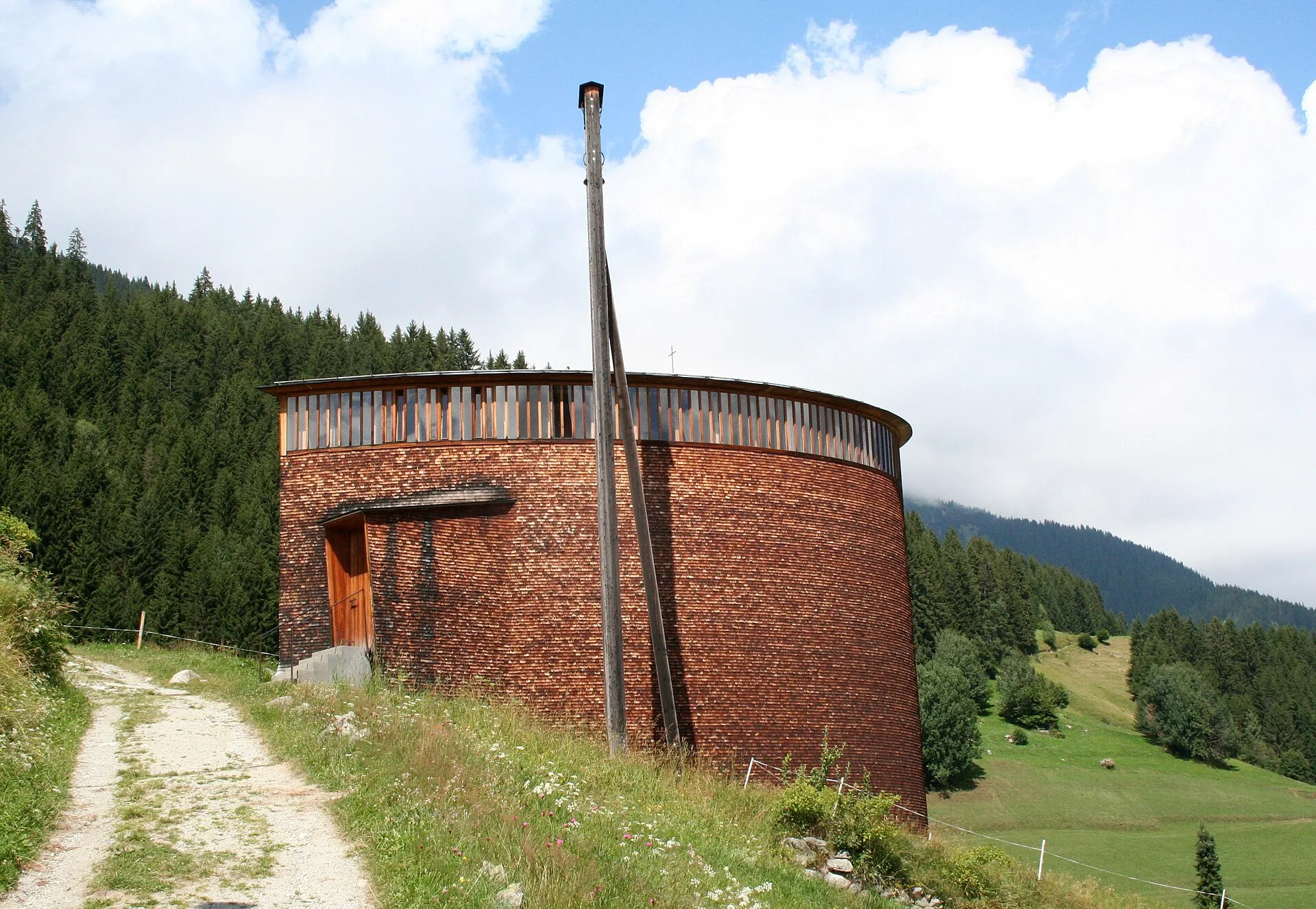 Small curved timber-shingled chapel with a slender bell post on a mountain path