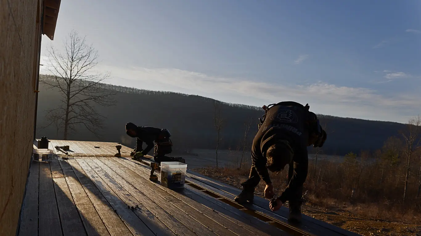 Two workers installing wood planks on a custom outdoor deck with a mountain landscape in the background during sunset.