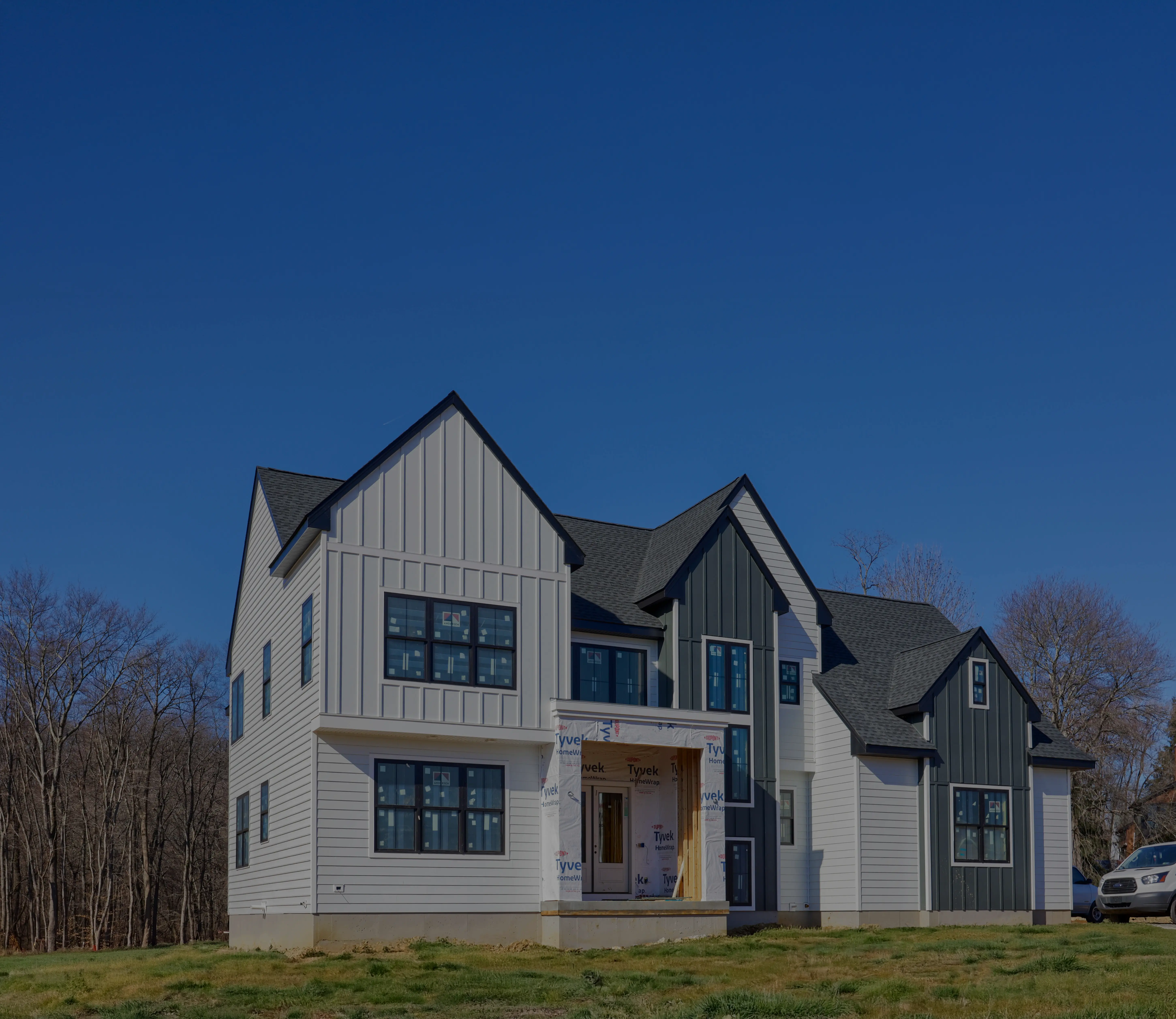 Two-story modern house under new construction with white and dark vertical siding, multiple windows, and new roofing being installed by Spruce Lane Construction.
