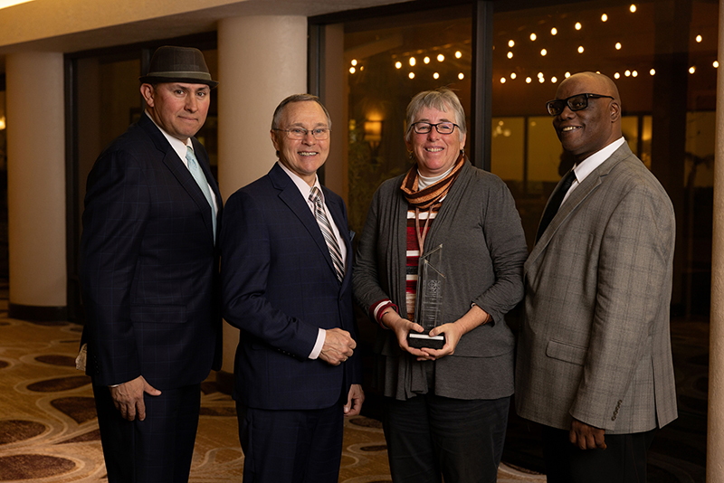 Four professionally dressed adults standing indoors, one woman holding a glass award.