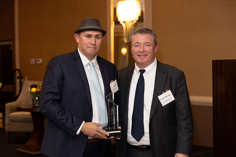 Two men in suits, one holding a glass award, standing indoors in a formal setting.