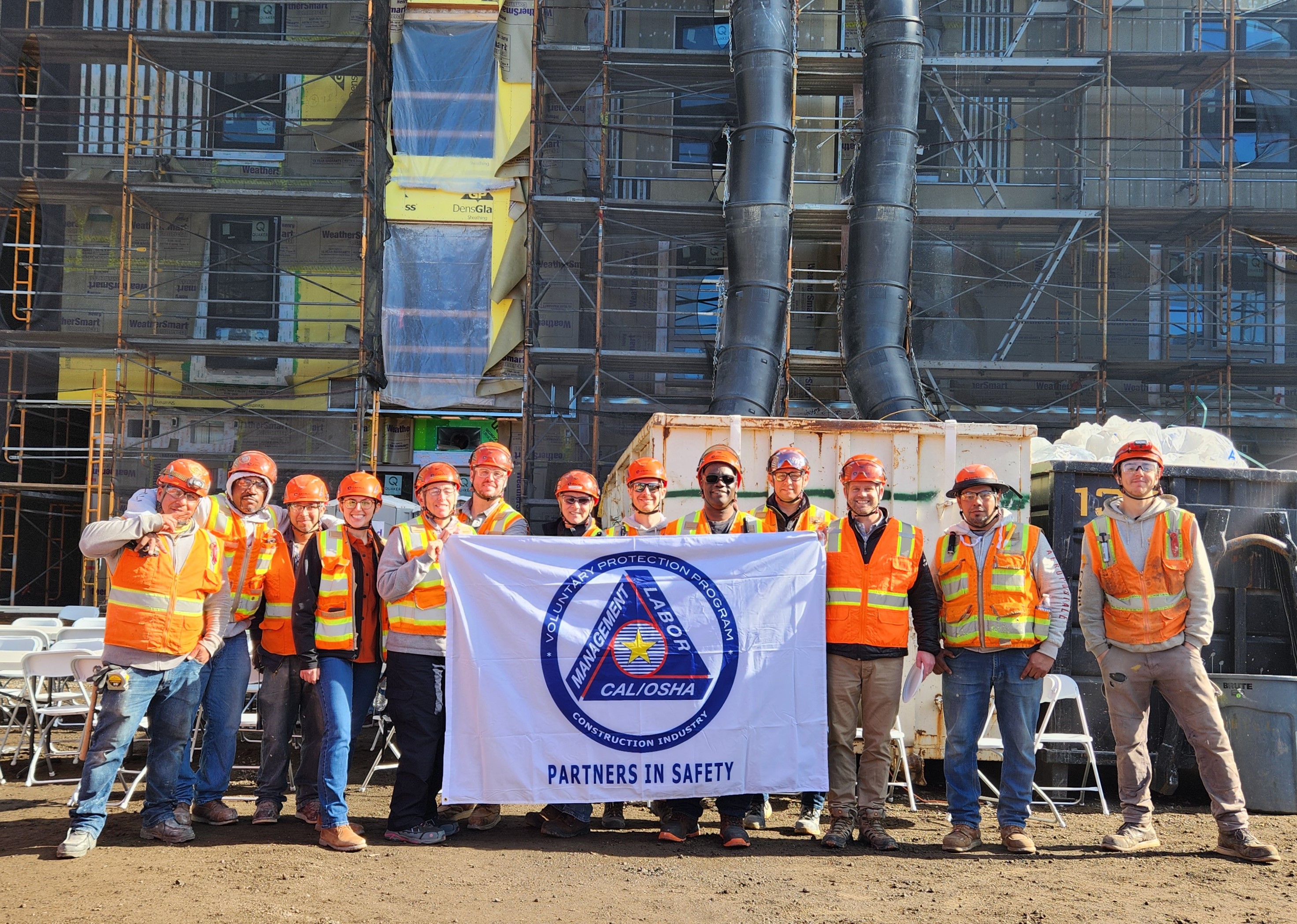 Group of construction workers in orange safety vests and helmets holding a CAL/OSHA Partners in Safety banner at a construction site.